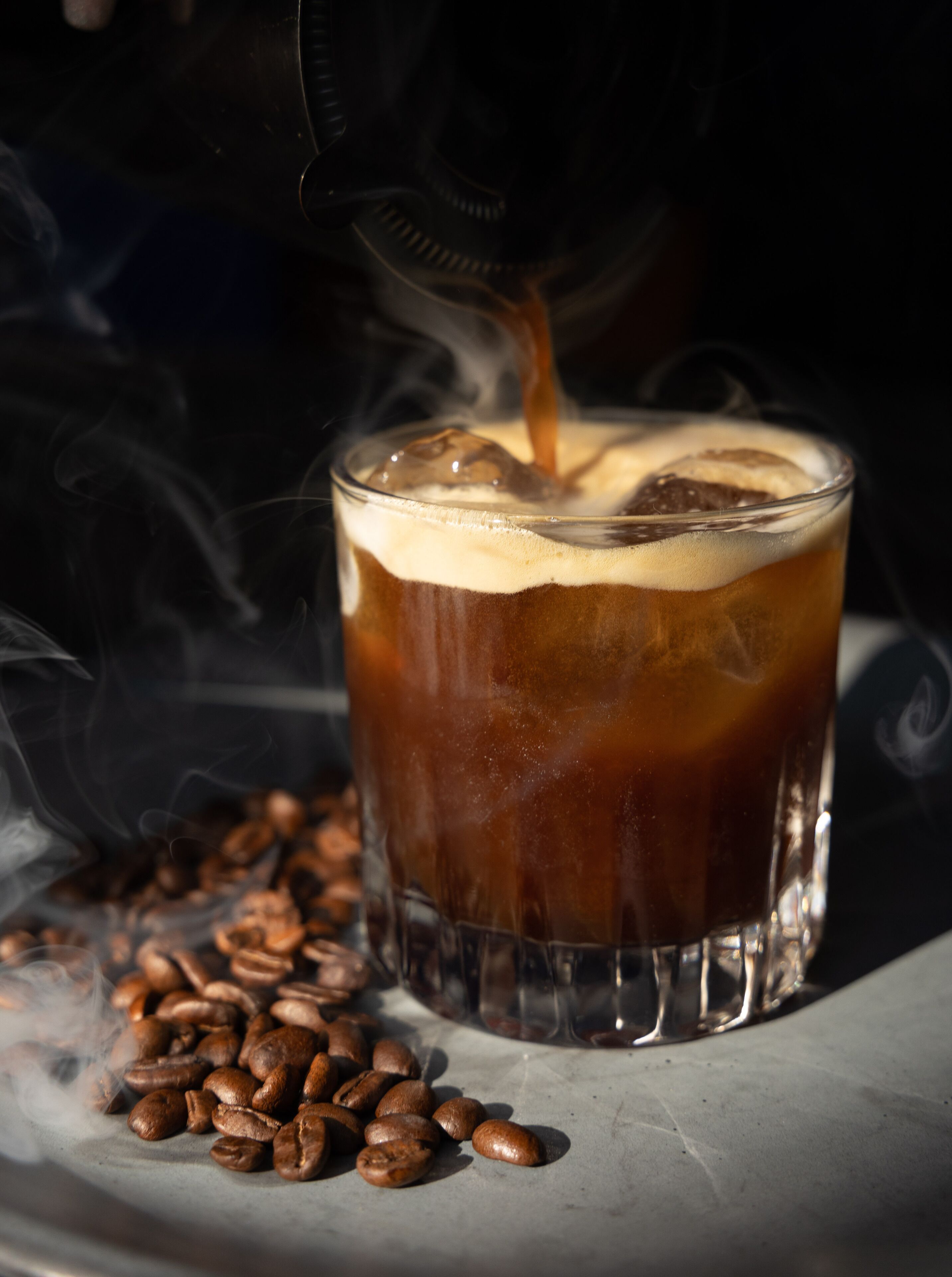 A glass of iced coffee with steam rising, surrounded by coffee beans on a marble surface.