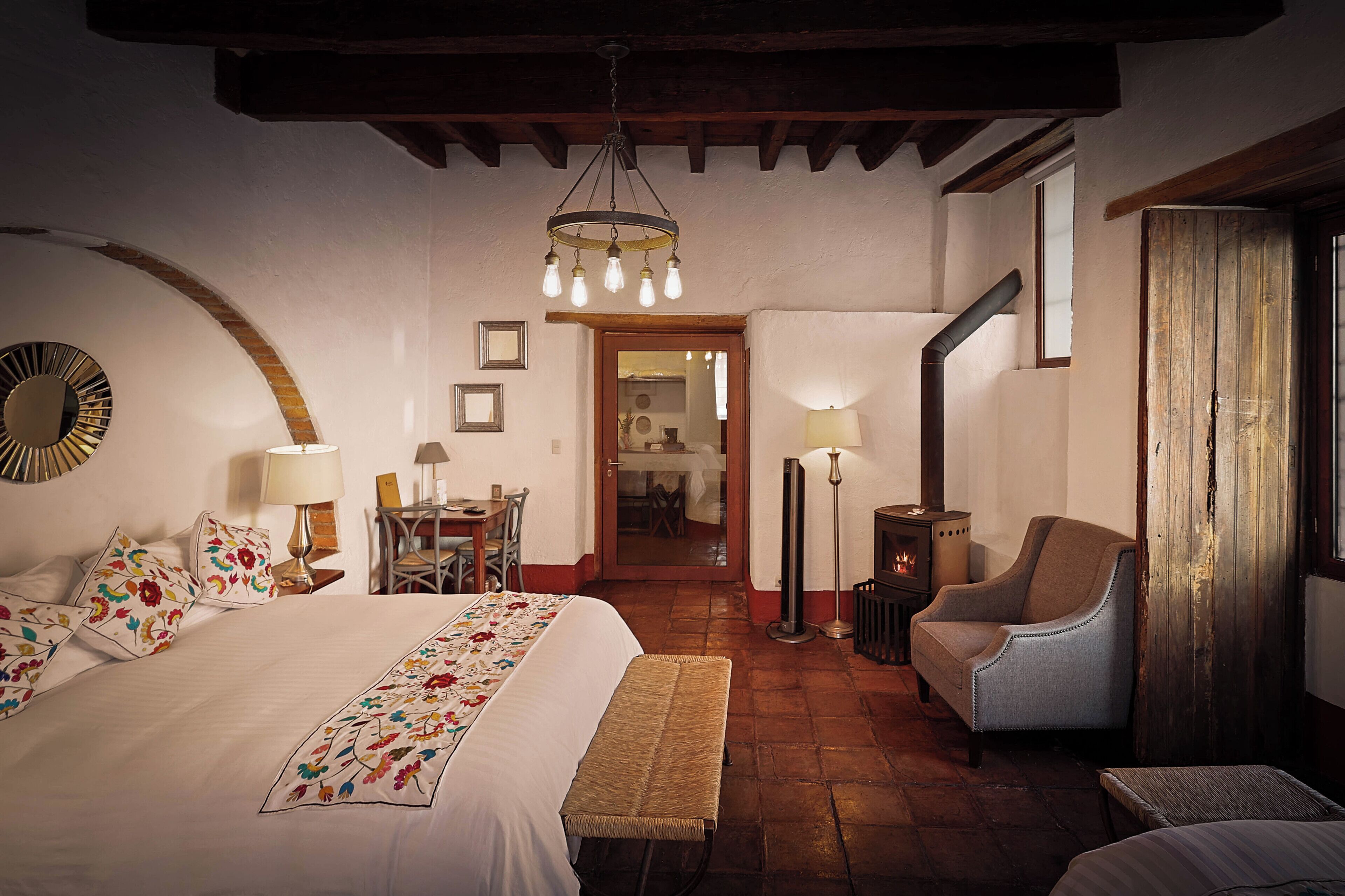 A cozy hotel room with a large bed, a bench, a chair, a table, and a fireplace.