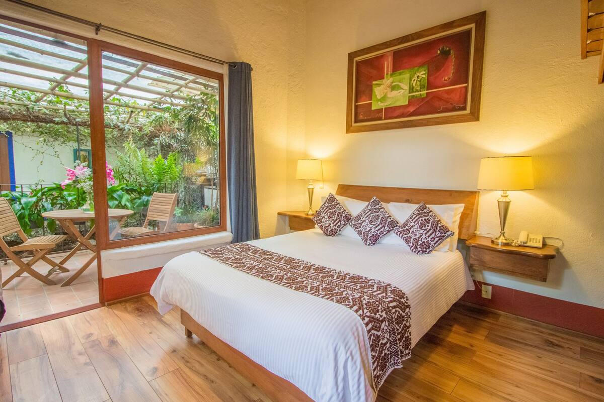 A bedroom with a large bed, two bedside lamps, a wooden headboard, a nightstand, and a window with a view of greenery.