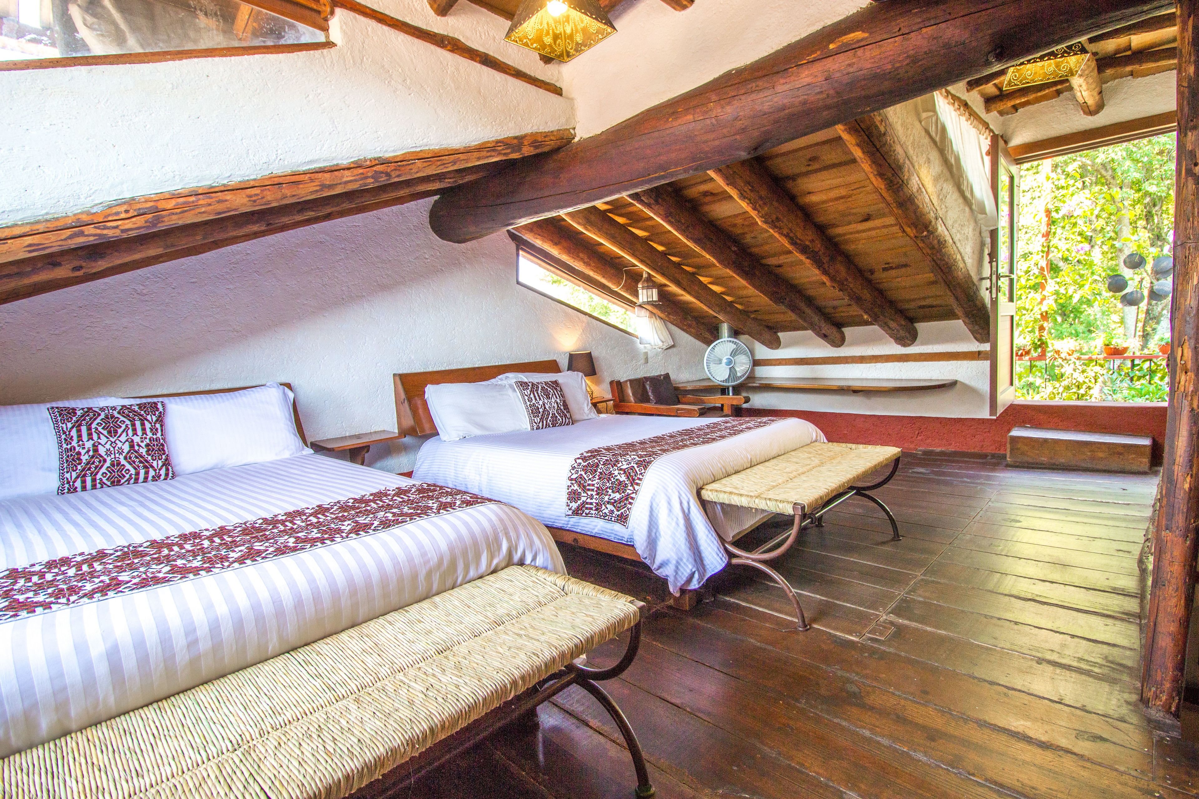 Habitación con dos camas, un banco, piso de madera y un ventanal con vistas a la vegetación.
