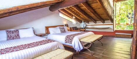 Habitación con dos camas, un banco, piso de madera y un ventanal con vistas a la vegetación.