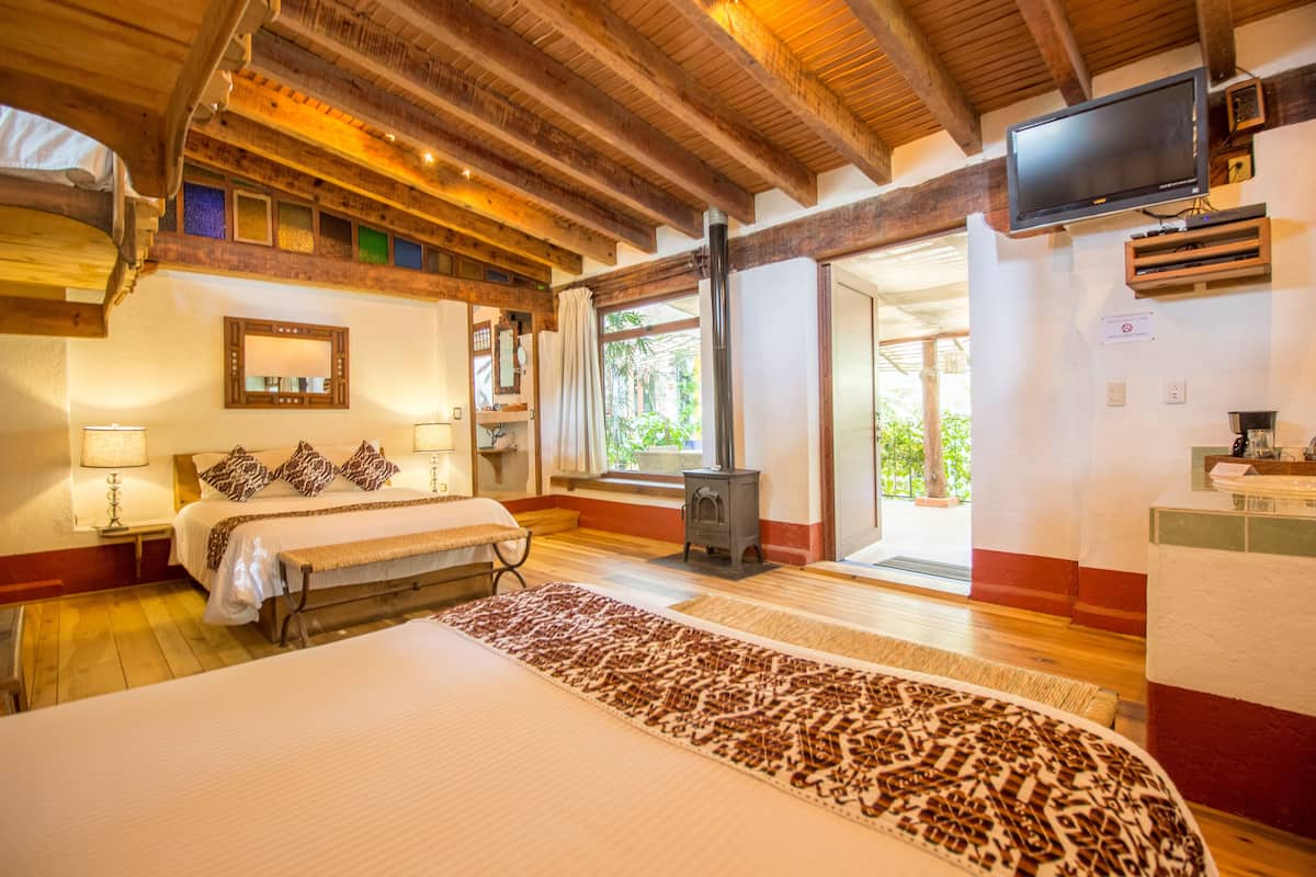 A spacious bedroom with a large bed, wooden ceiling beams, a television, and a window with a view of greenery.