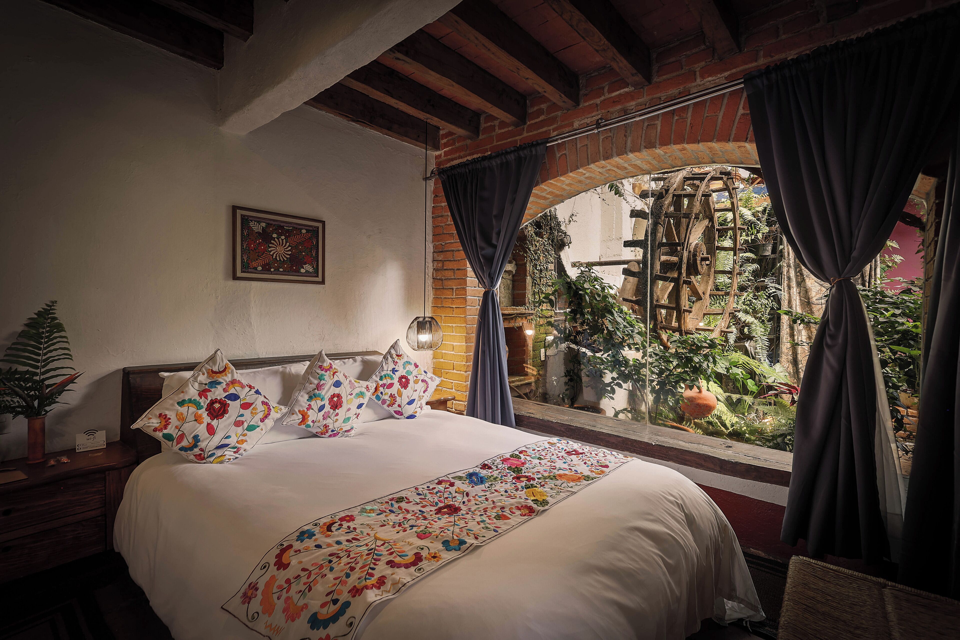 A bedroom with a large bed, a window with curtains, a potted plant, and a rustic brick wall.