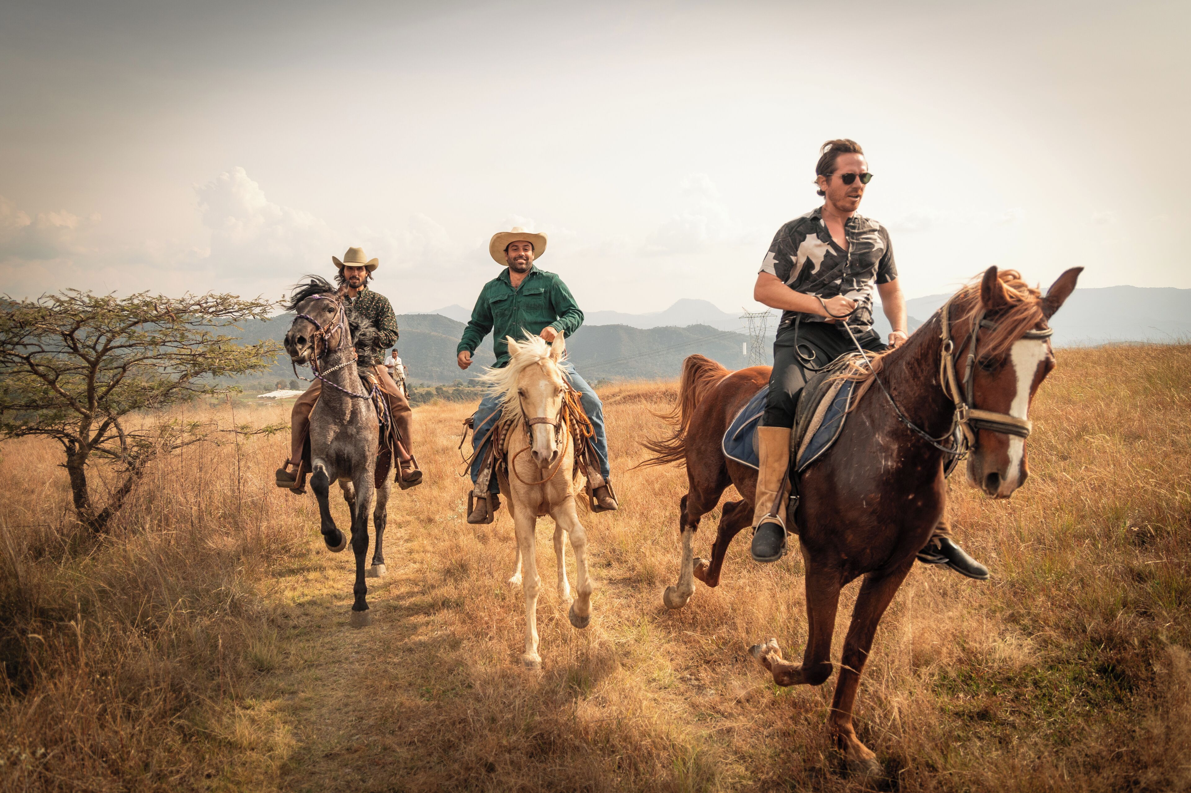 Drie personen te paard in een grasveld, met bergen op de achtergrond.
