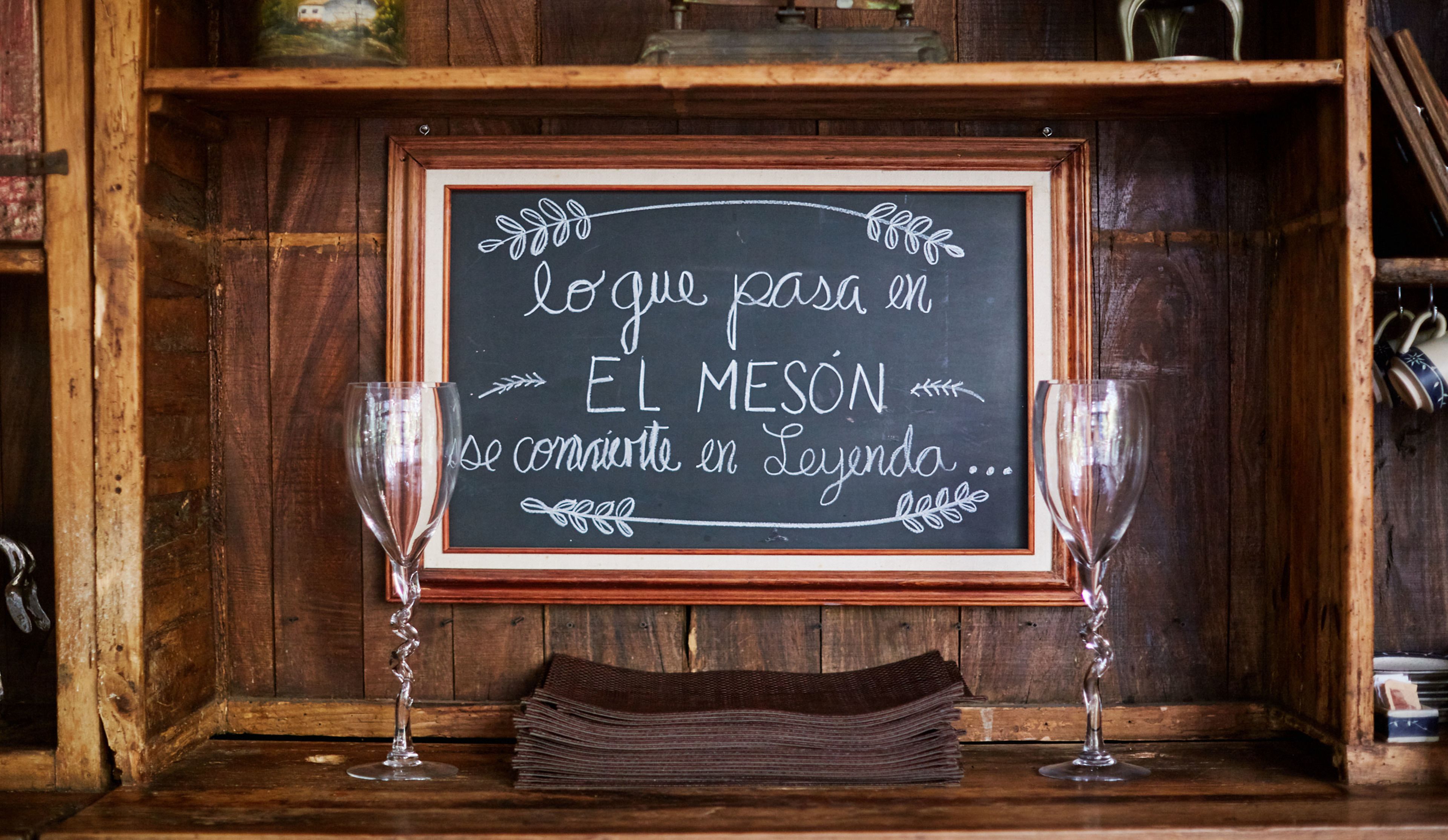 Una lavagna in legno con un messaggio in spagnolo, due bicchieri da vino e una pila di menu su un ripiano.
