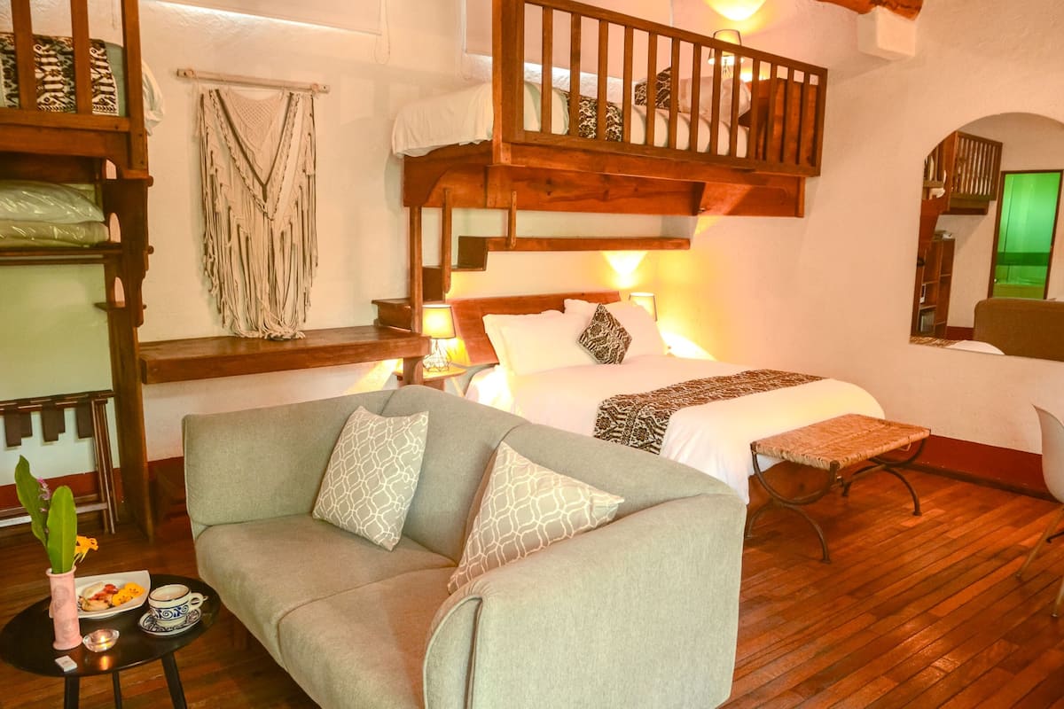 A cozy room with a bunk bed, a sofa, a bedside table with a lamp, and a small table with a plant and a plate of food.
