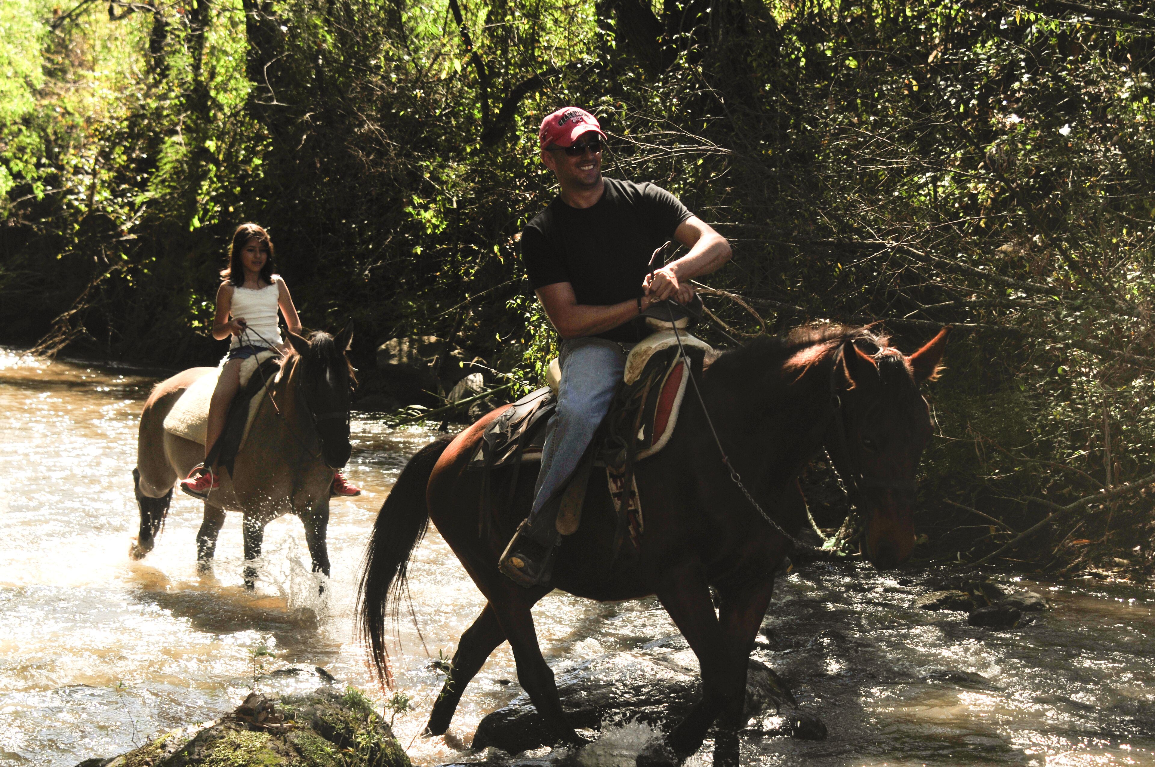 Two people riding horses through a shallow stream in a forested area.