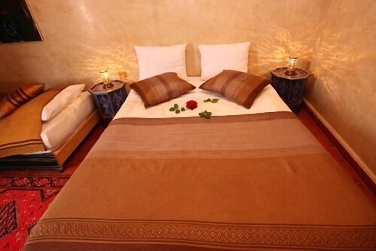A neatly made bed with a brown bedspread and two pillows, a bedside table with a lamp, and a red rose on the bed.