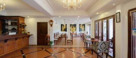 Un hall de hotel con mostrador de recepción, una araña, un área de comedor con mesas y sillas, y un reloj en la pared.