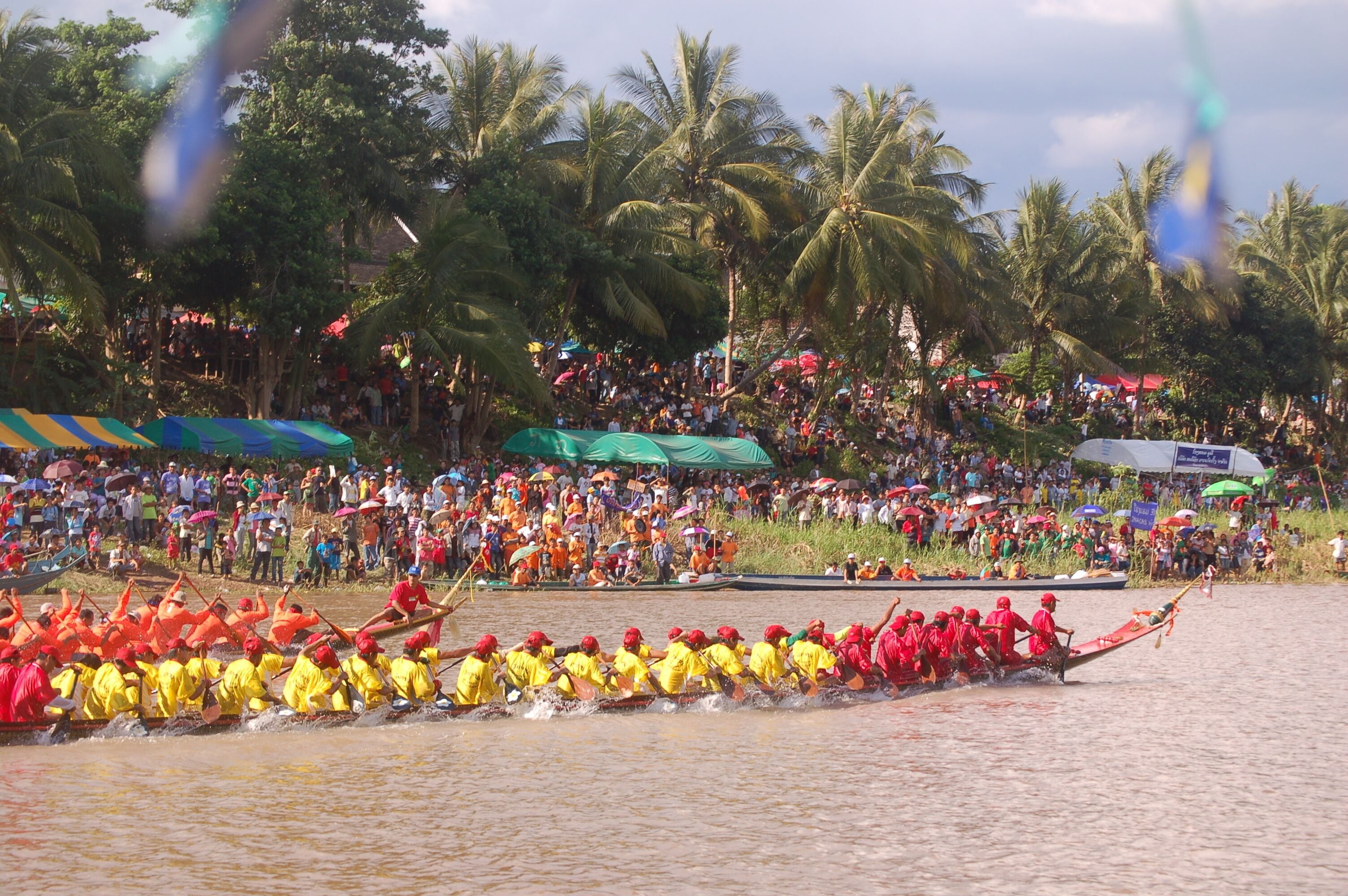 A dragon boat race with teams in yellow and red uniforms, surrounded by a large crowd and palm trees.