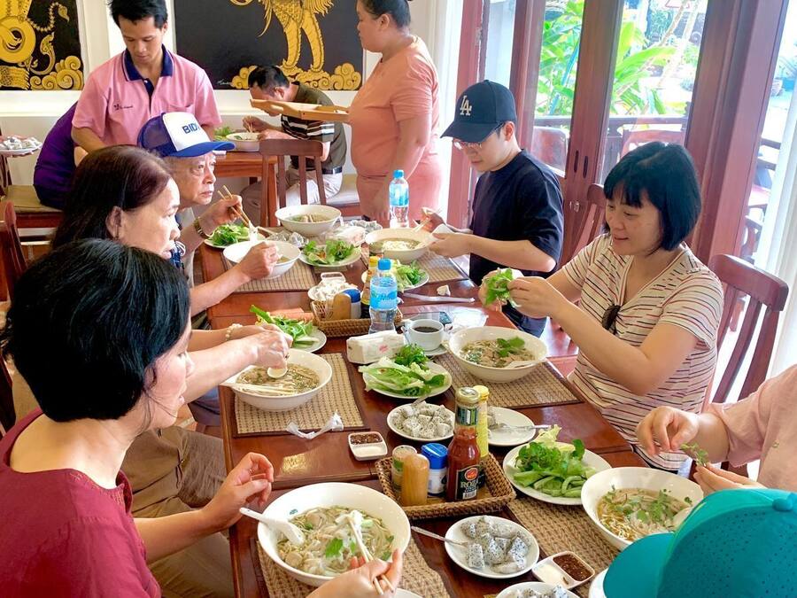Un grupo de personas cenando en un restaurante, con una mesa grande llena de diferentes platos, incluyendo tazones de sopa y bandejas de comida.