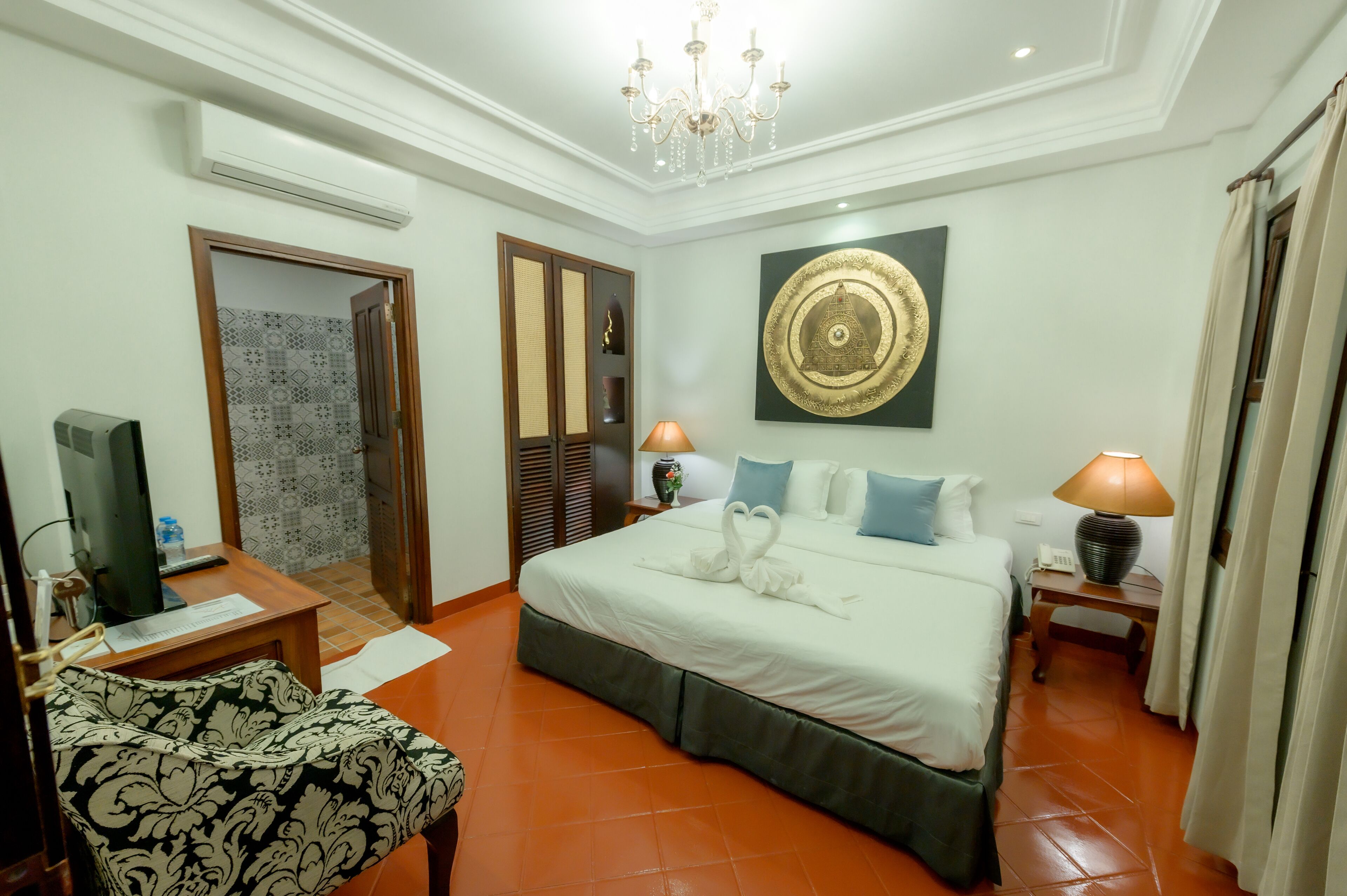 A hotel room with a bed, a desk with a computer, a chair, a wall-mounted TV, a decorative wall piece, and a chandelier.