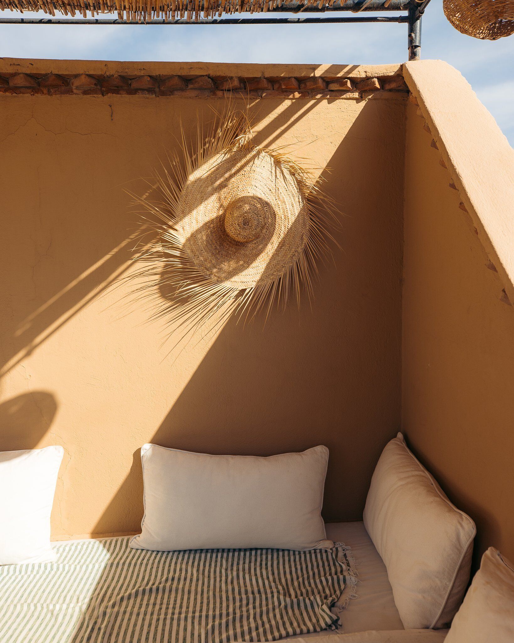 A small, enclosed balcony with a woven sunshade, a bed with white pillows, and a striped blanket.