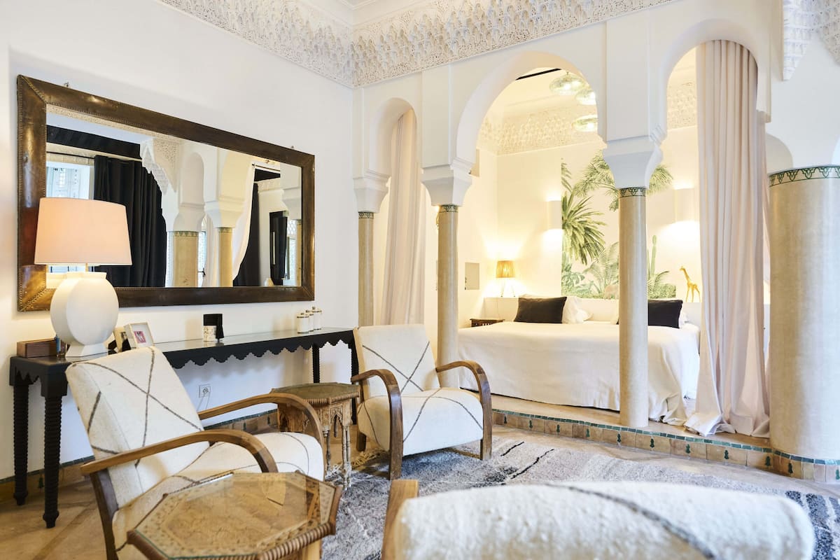 A hotel room with a large mirror, a bed, a sitting area with chairs, and a view through an archway.