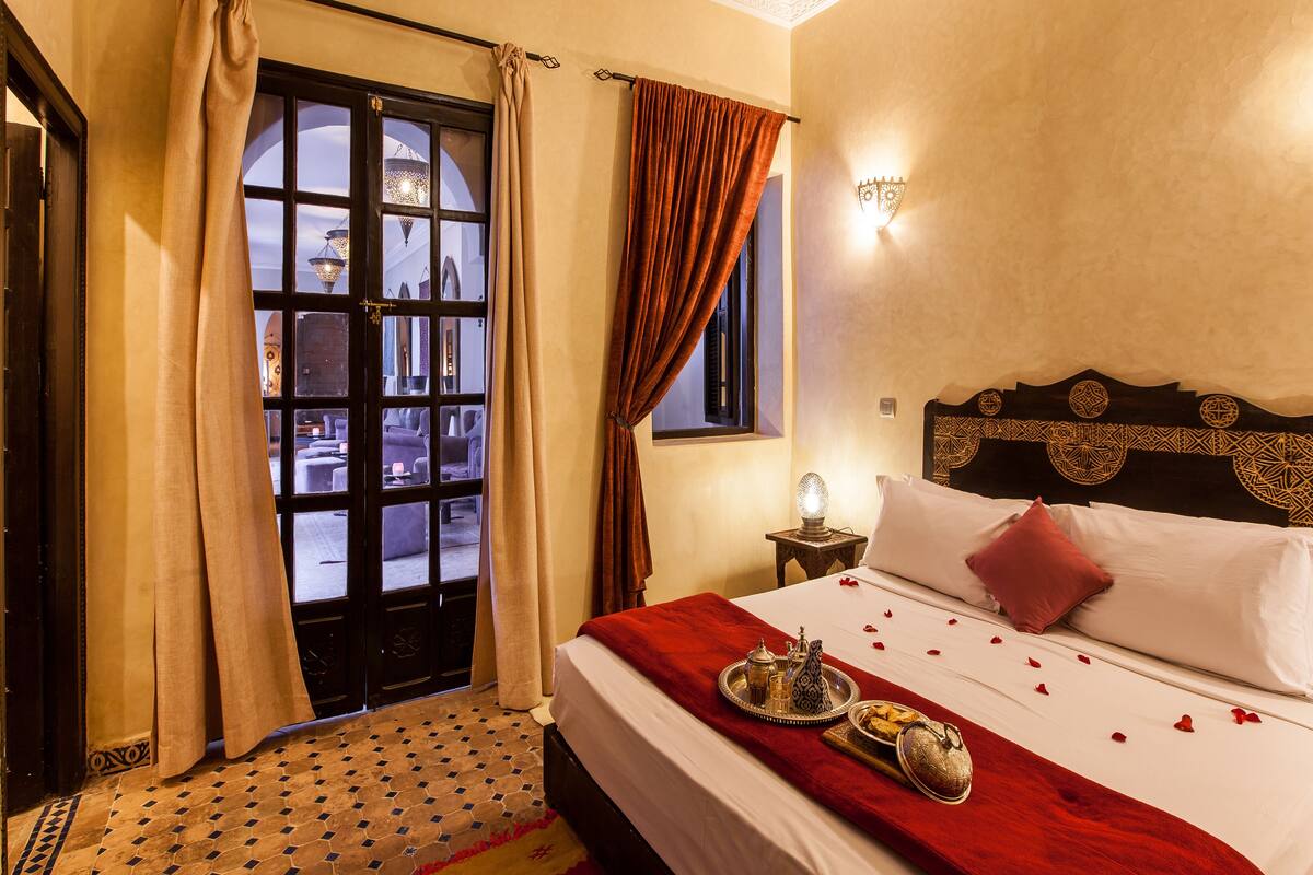 A hotel room with a large bed, a nightstand with a lamp, and a view through a window with curtains.