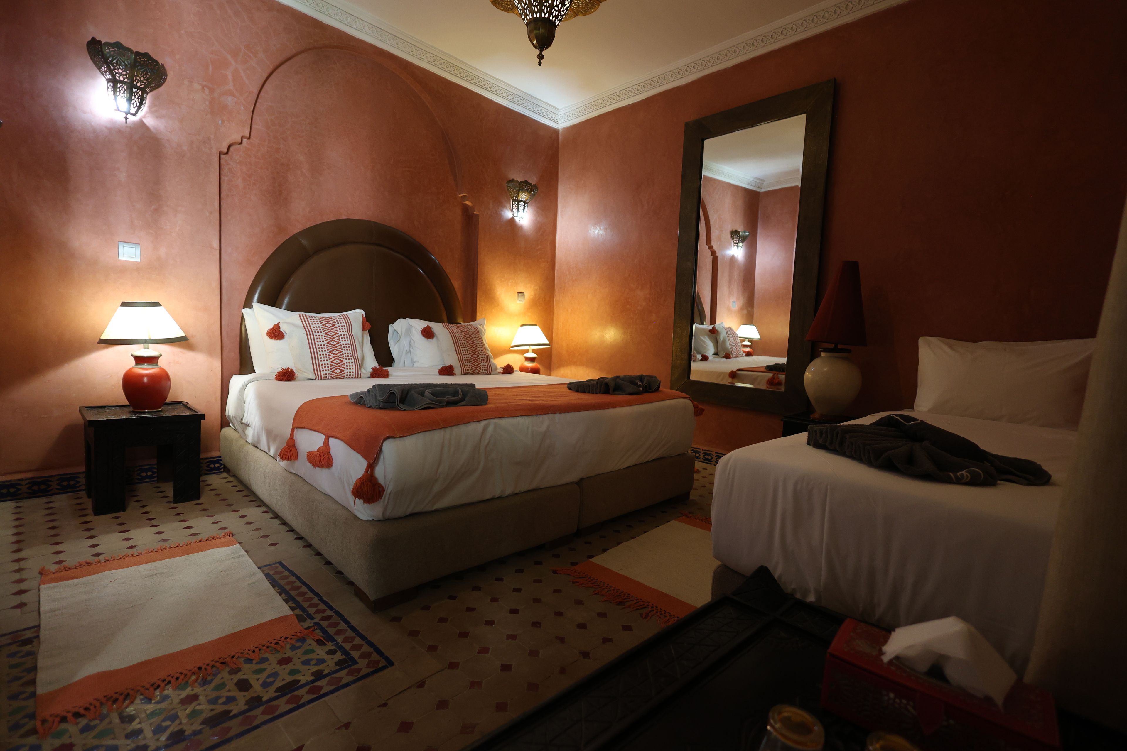 A hotel room with a large bed, a smaller bed, a nightstand with a lamp, a mirror, and a patterned rug.