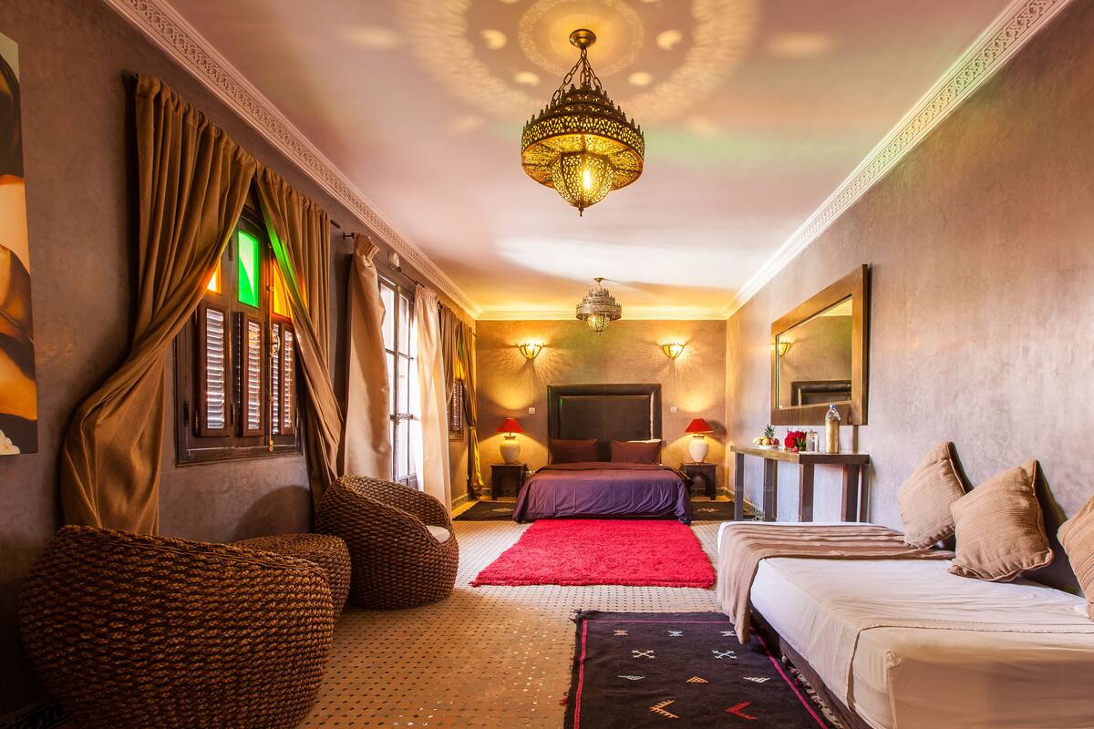 A hotel room with a large bed, a sitting area with a sofa and armchairs, a red rug, and a chandelier.