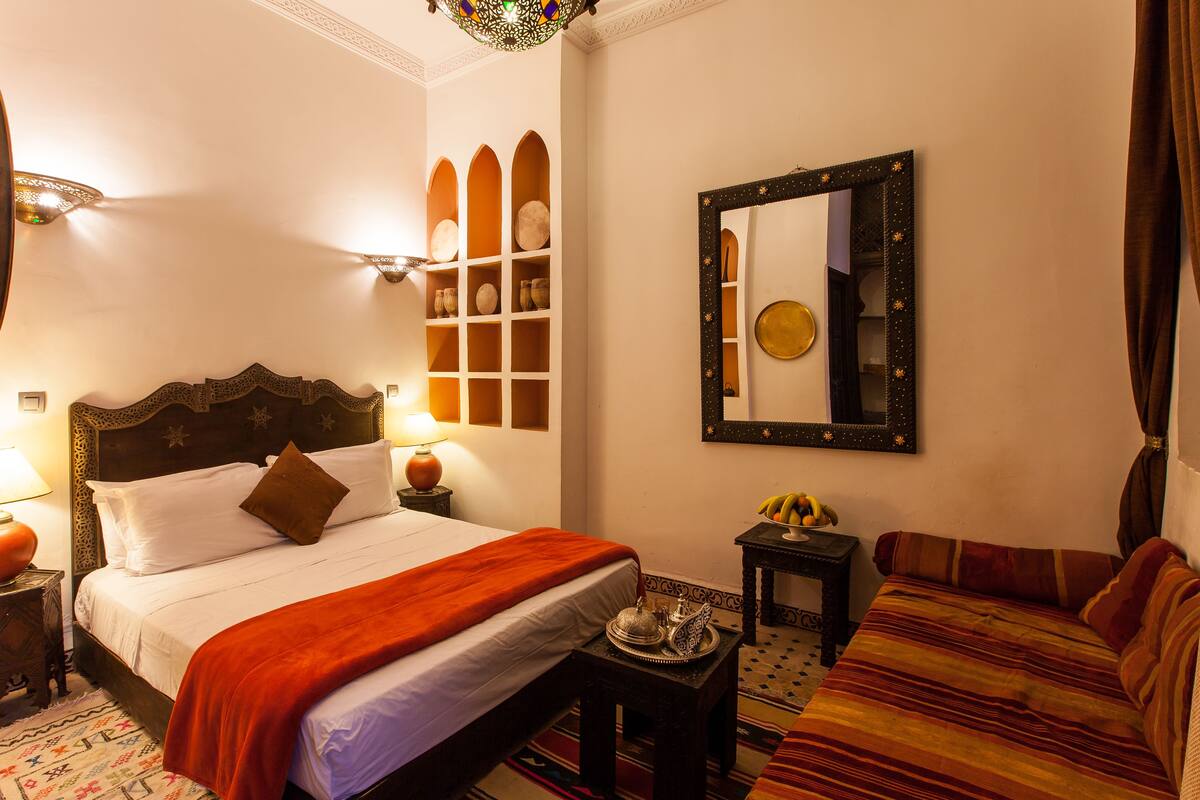 A well-lit bedroom with a large bed, a nightstand, a small table with a decorative item, a sofa, and a wall-mounted mirror.
