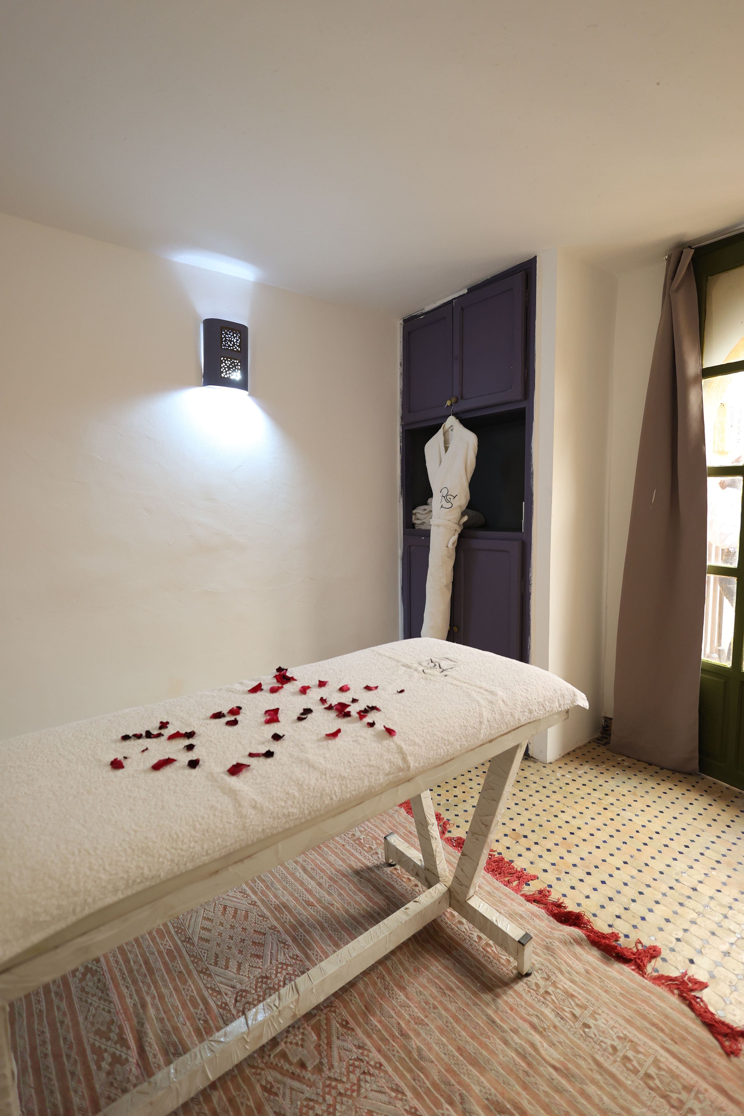 A room with a bed, a small table with rose petals, a robe hanging on a door, and a patterned rug.