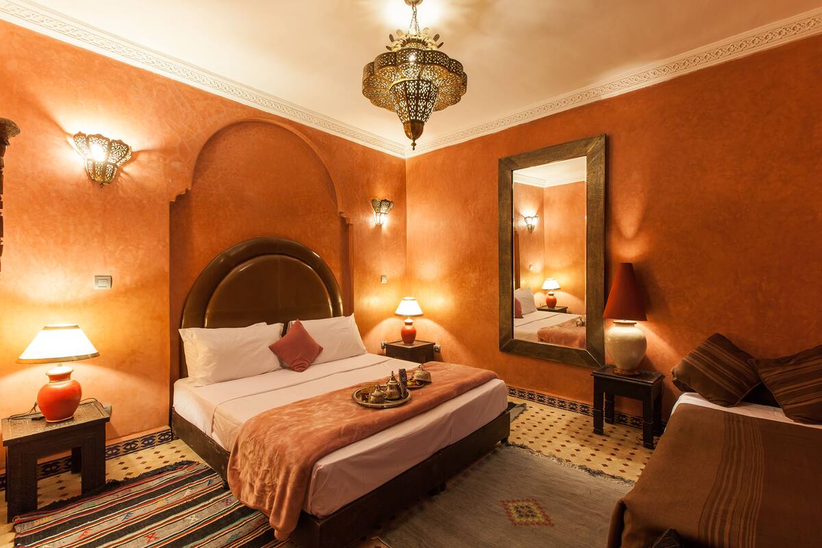 A hotel room with a large bed, two bedside lamps, a chandelier, a mirror, and a patterned rug.