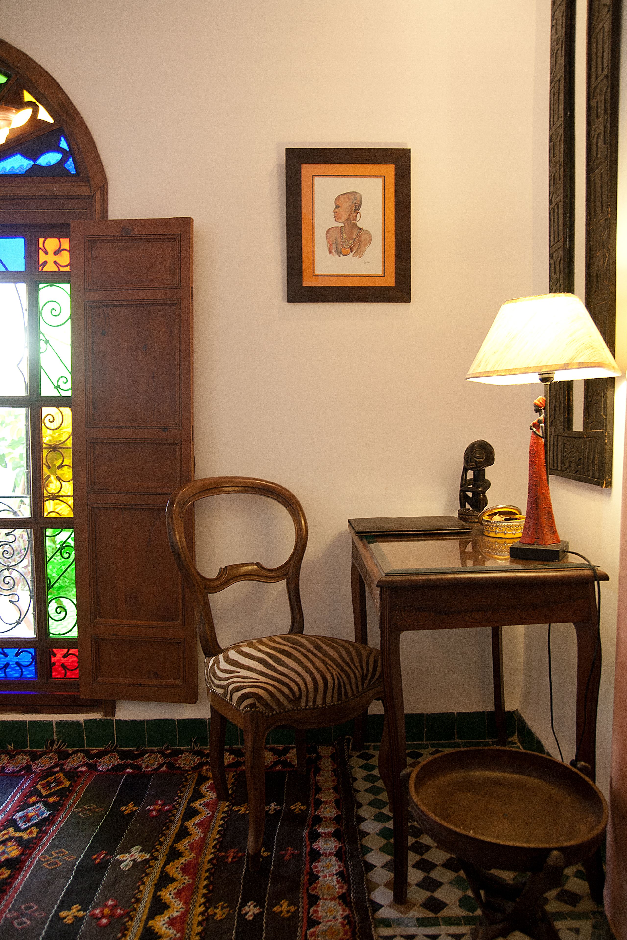 A room with a wooden chair, a table with a lamp, a framed picture on the wall, and a stained glass window.
