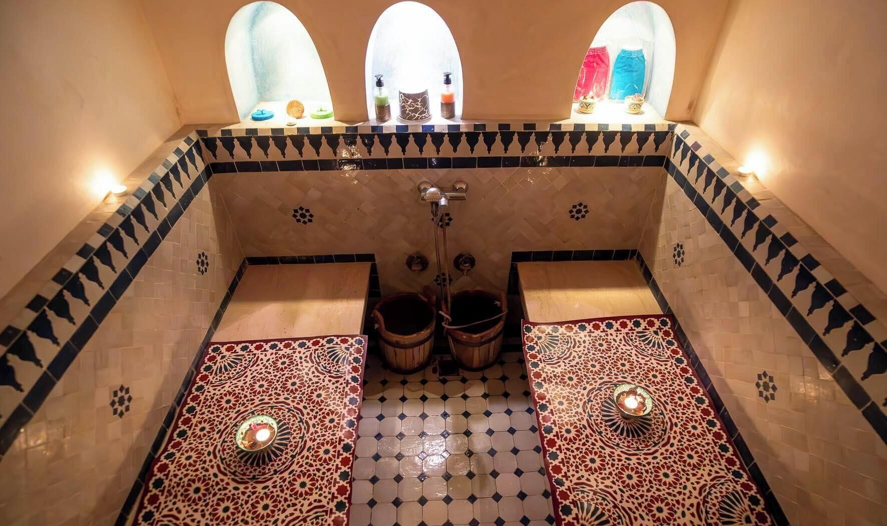 Un baño de estilo marroquí tradicional, con suelo de baldosas, dos cubos de madera, un lavamanos central y tres ventanas de arco con toallas de colores.