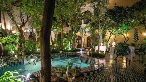 Une piscine entourée d’une végétation luxuriante, un sol carrelé et une table avec des chaises.