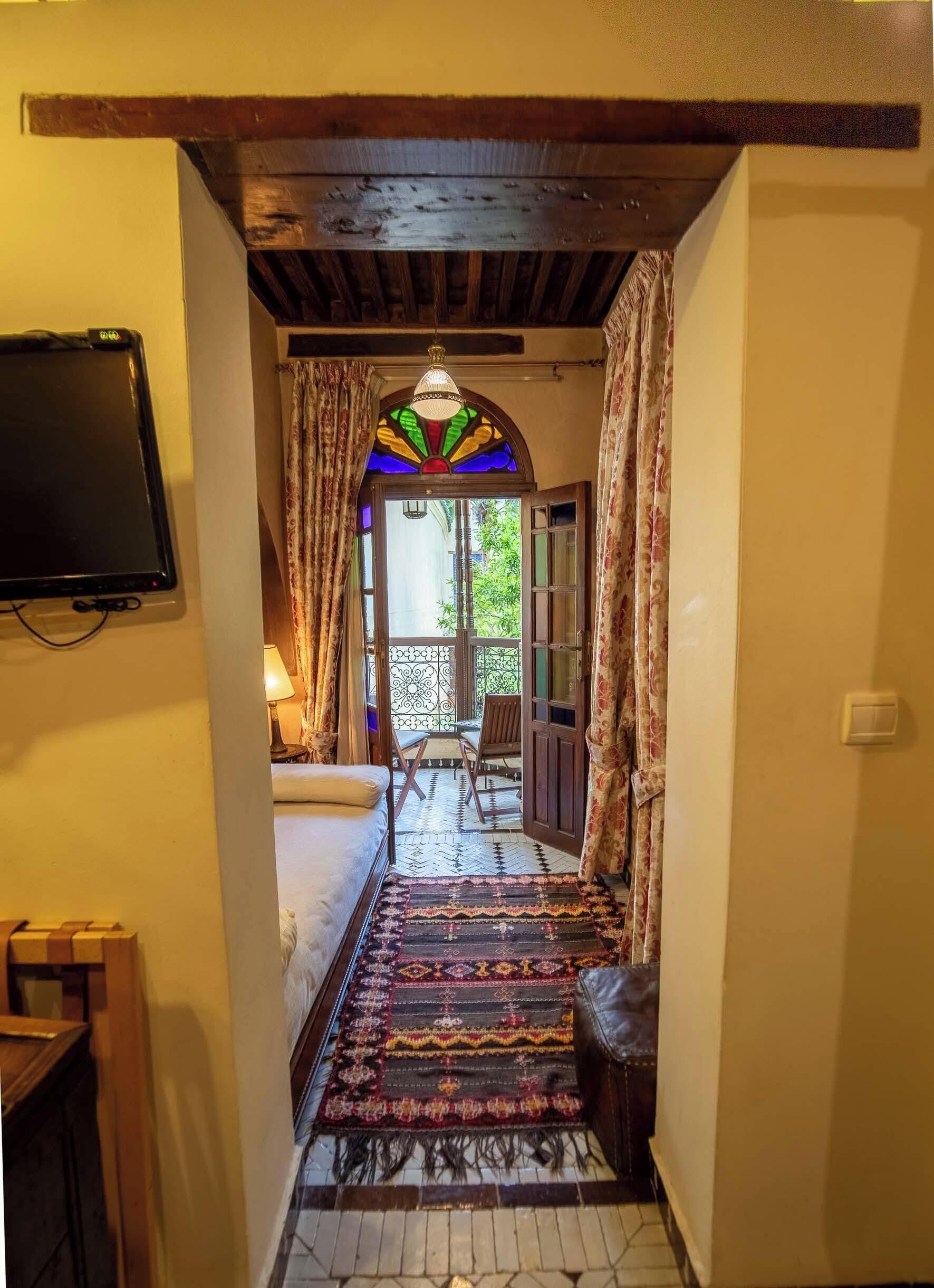 A hallway leading to a balcony with a patterned rug, a bed, and a television mounted on the wall.