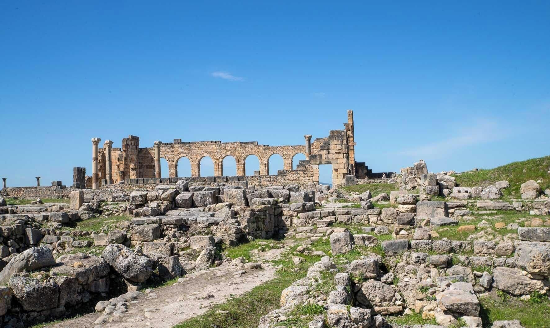 Ancient ruins with stone columns and arches, surrounded by scattered stones and grass.