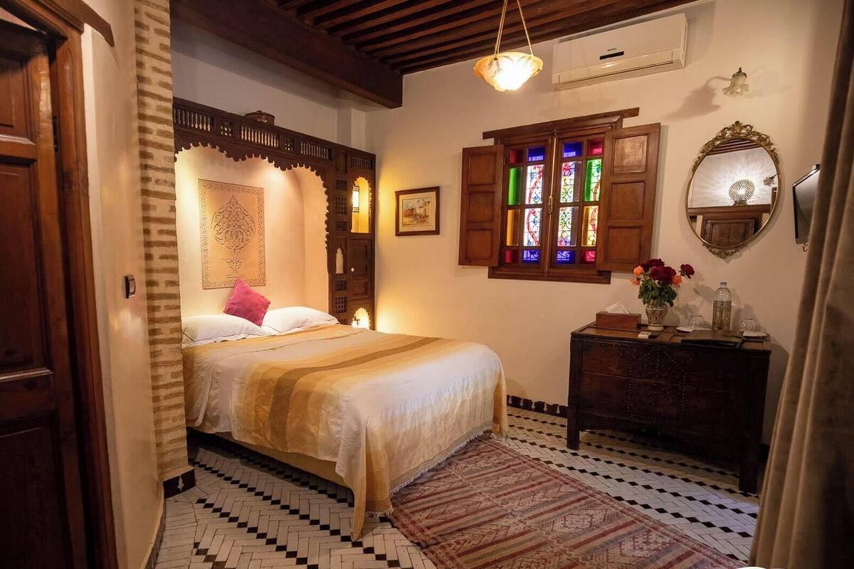 Ein Schlafzimmer mit einem Bett, einem Schminktisch, einem Spiegel, einem Fenster mit Buntglas und einem gemusterten Teppich.