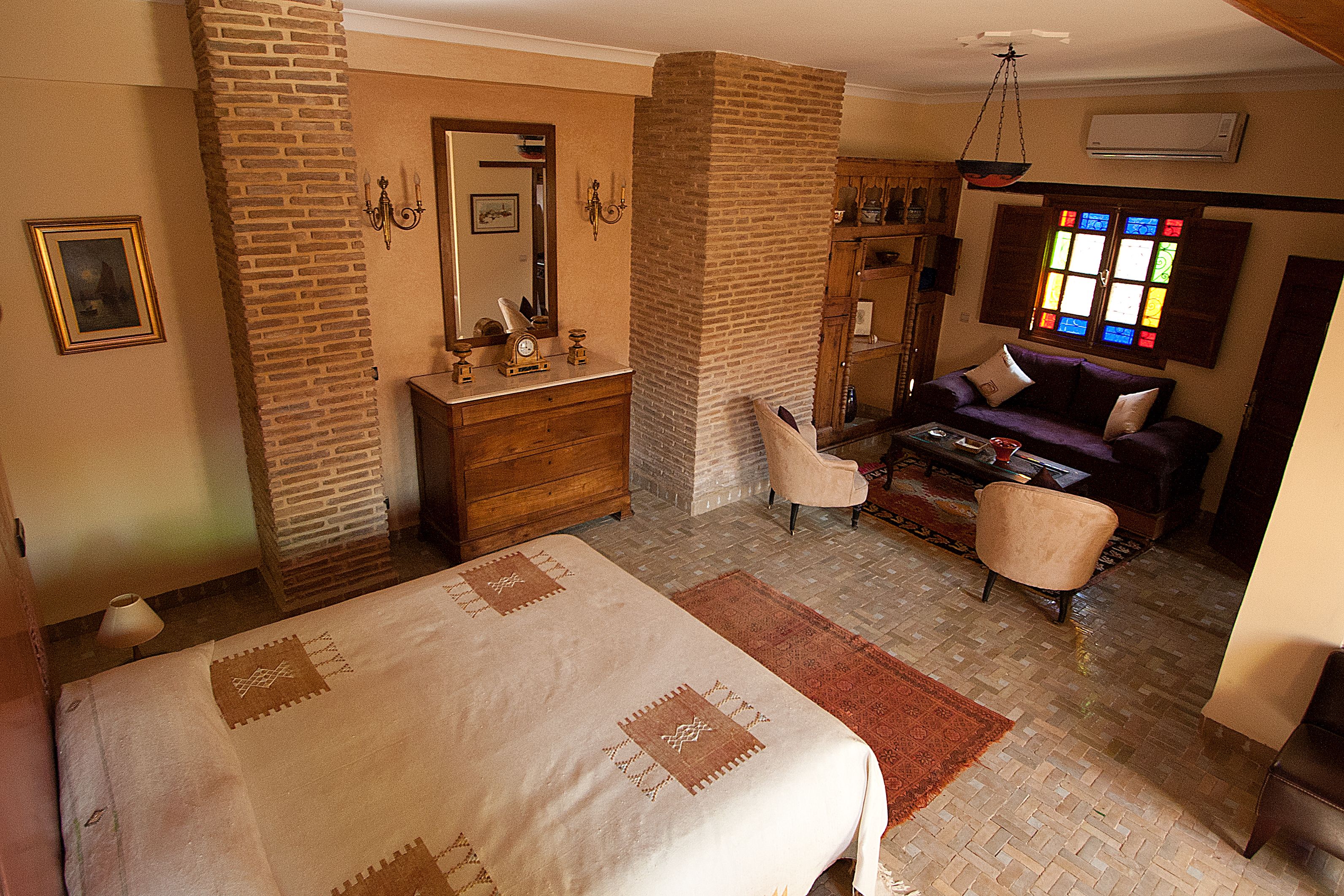 A bedroom with a large bed, a dresser, a vanity, a chair, and a window with stained glass.