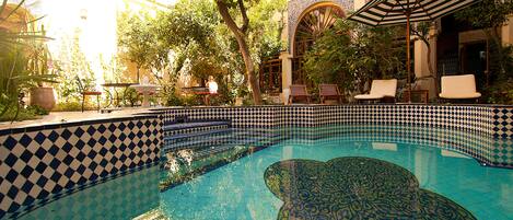 Una piscina con azulejos estampados, rodeada de sillones de descanso y una mesa, con un edificio con diseño de baldosas a cuadros al fondo.