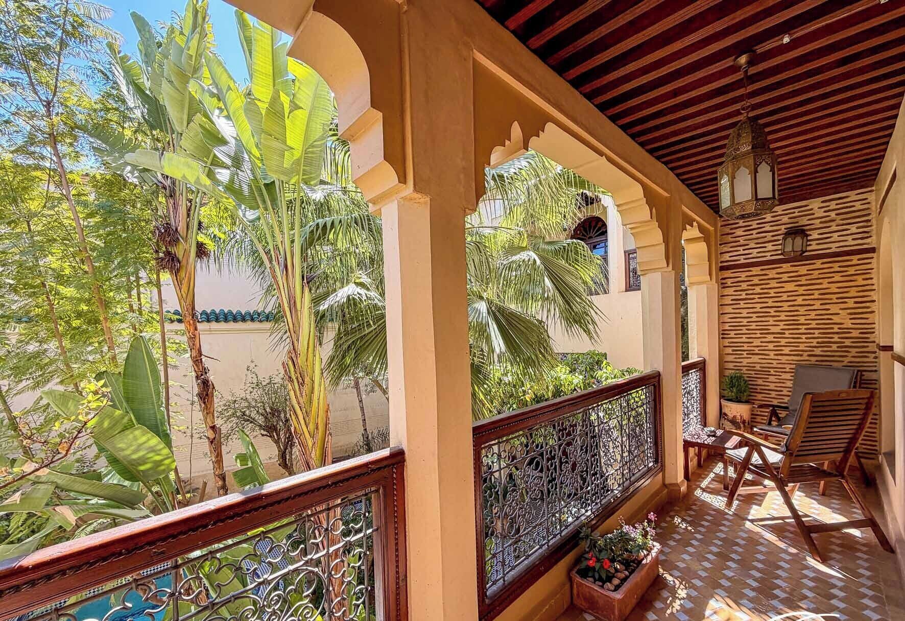 A covered balcony with a view of tropical vegetation, a tiled floor, and a seating area with chairs and a table.