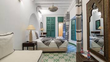 Habitación de hotel con cama, dos mesitas de noche, espejo, candelabro, puertas verdes y baldosas de suelo con diseño.