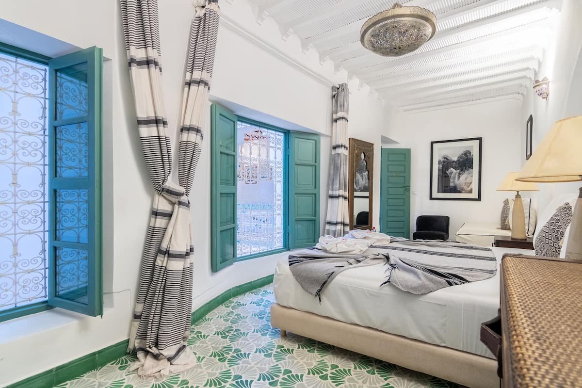 Een kamer met een bed, een stoel, een bureau, een raam met gordijnen en een kroonluchter.