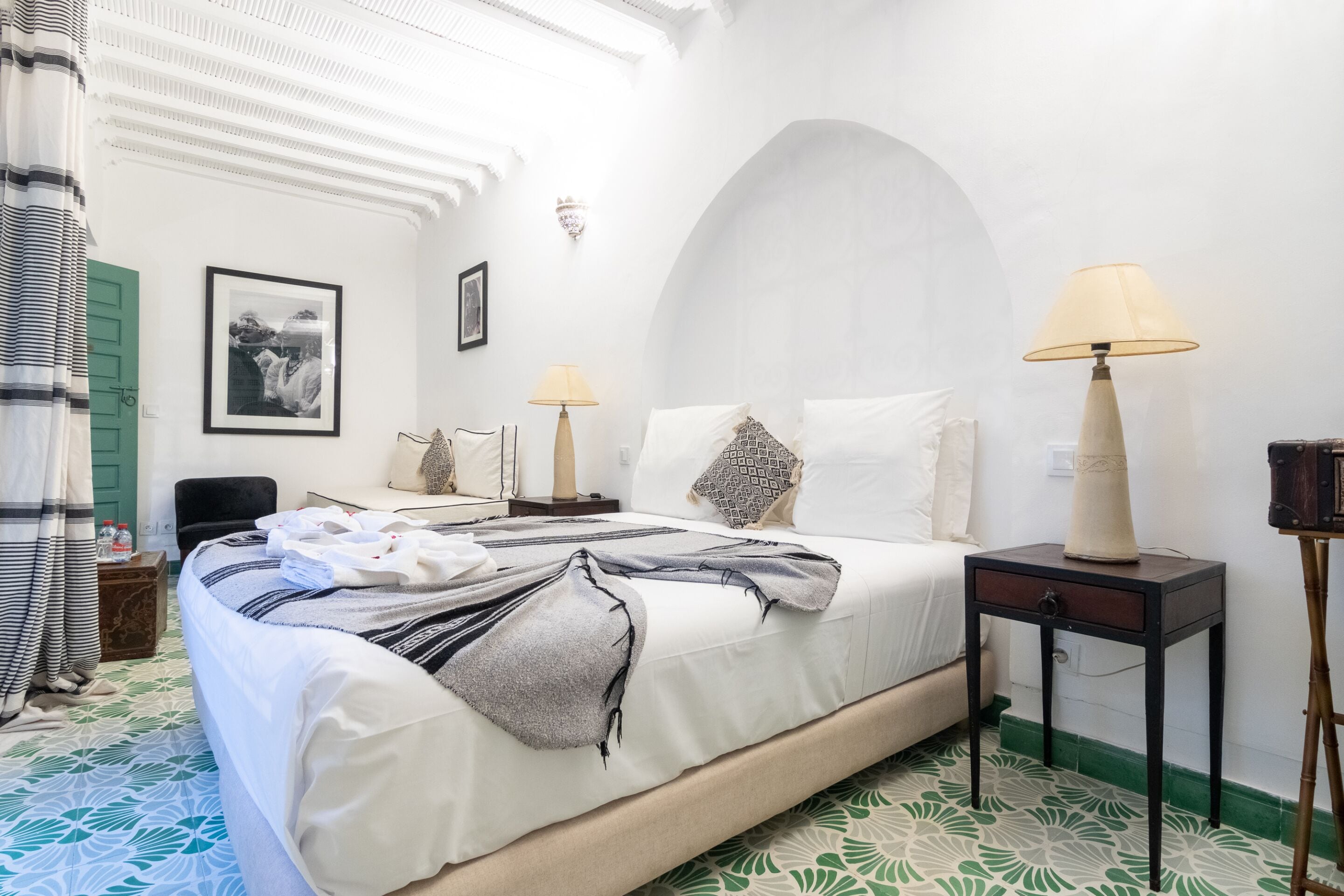 A hotel room with a bed, two bedside tables, a chair, a green door, and a framed picture on the wall.