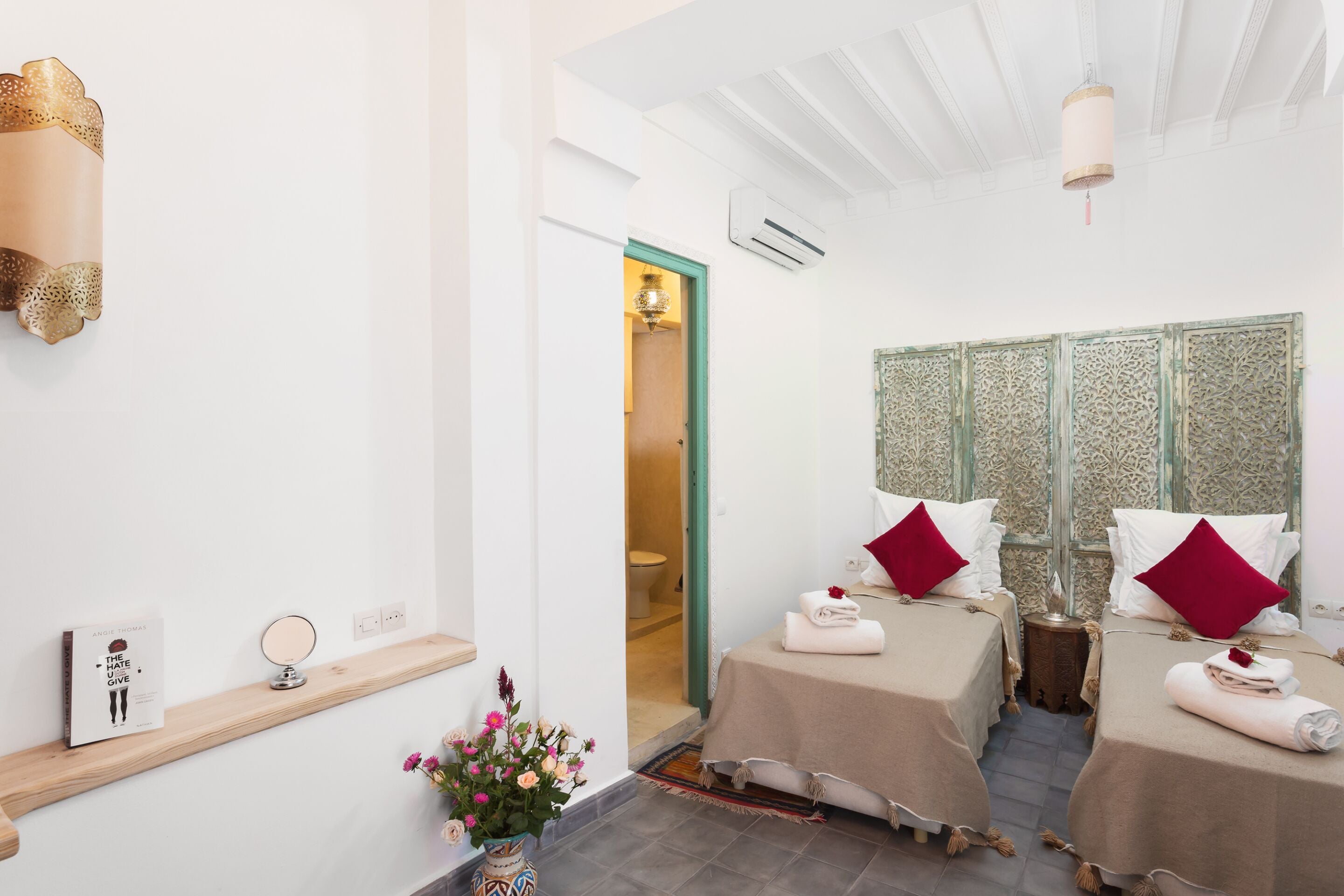 Habitación de hotel con dos camas, un espejo, un jarrón con flores, una estantería con un libro y un panel decorativo en la pared.