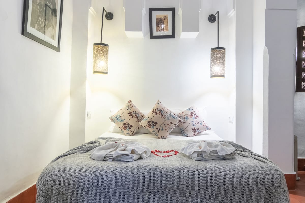 Een netjes opgemaakt bed met bloemenkussens, een beddekleed met een patroon en een rode hartdecoratie. Boven het bed hangen twee wandlampen en er zijn afbeeldingen in lijsten.