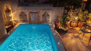 Una piscina rectangular con agua cristalina, rodeada por un patio con baldosas, plantas en macetas y mobiliario exterior.