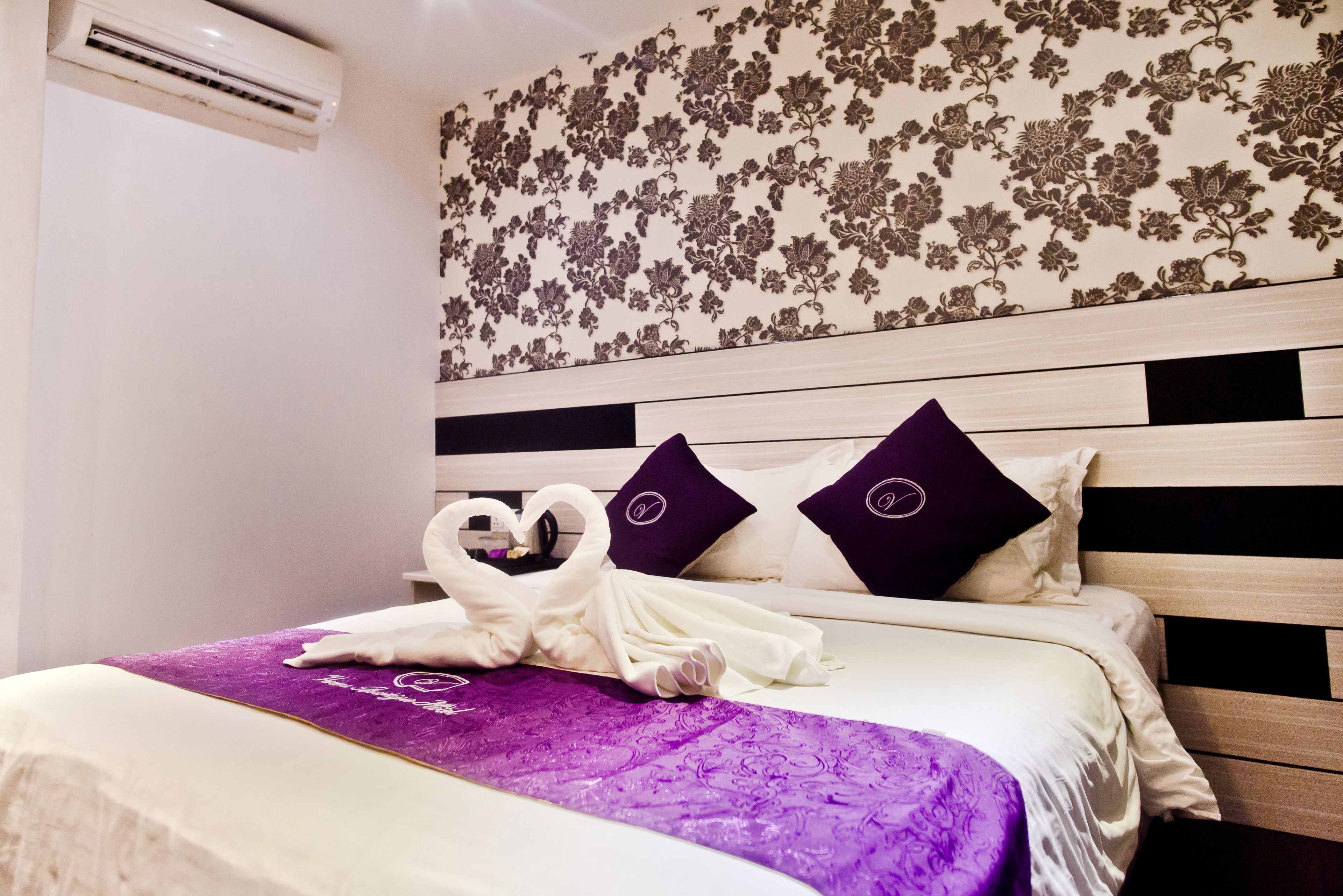 A hotel room with a bed, a purple bedspread, and a floral wallpaper design.
