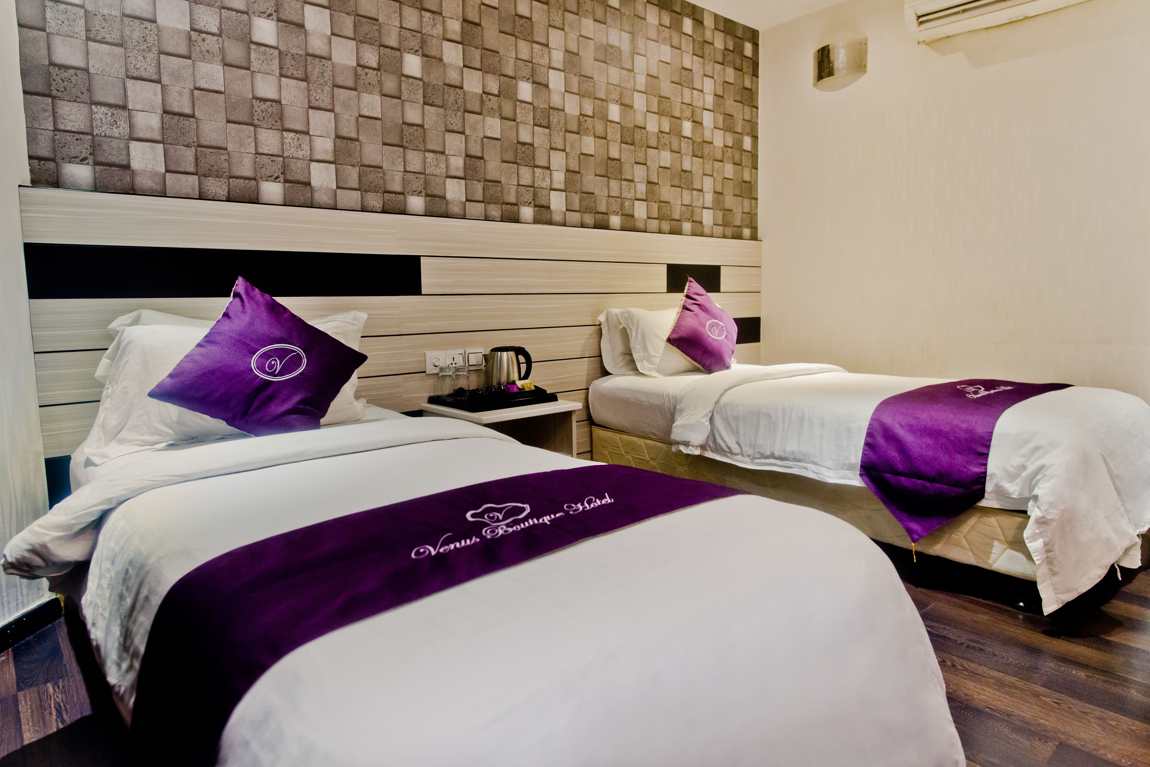 A hotel room with two beds, each with a purple bedspread and matching pillow, a wooden headboard, and a brick-patterned wall.