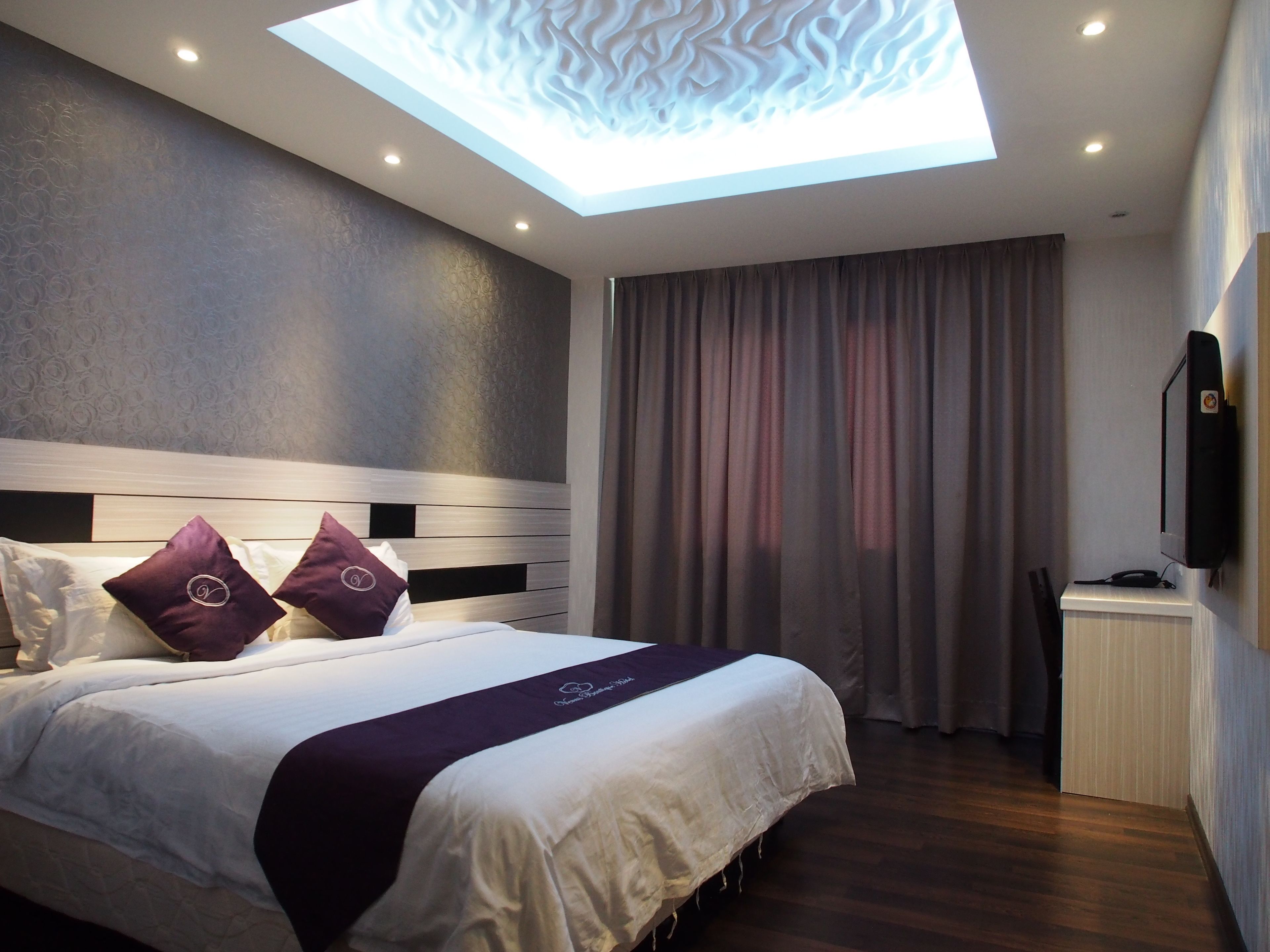A modern hotel room with a skylight, a bed with white bedding and purple accents, a desk with a flat-screen TV, and a wooden floor.