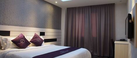 A modern hotel room with a skylight, a bed with white bedding and purple accents, a desk with a flat-screen TV, and a wooden floor.