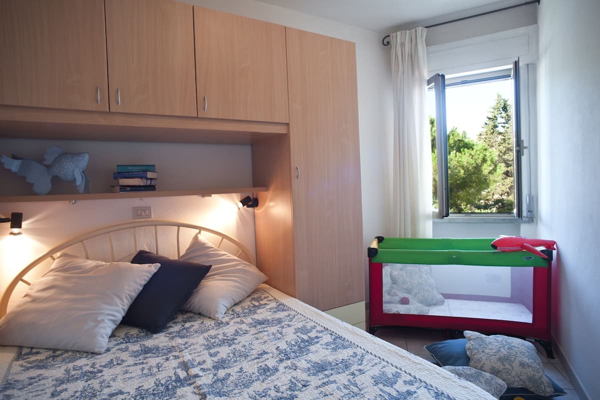 Ein Schlafzimmer mit Bett, Nachttisch, Fenster mit Vorhängen und einem kleinen, grün-roten Reisebett.