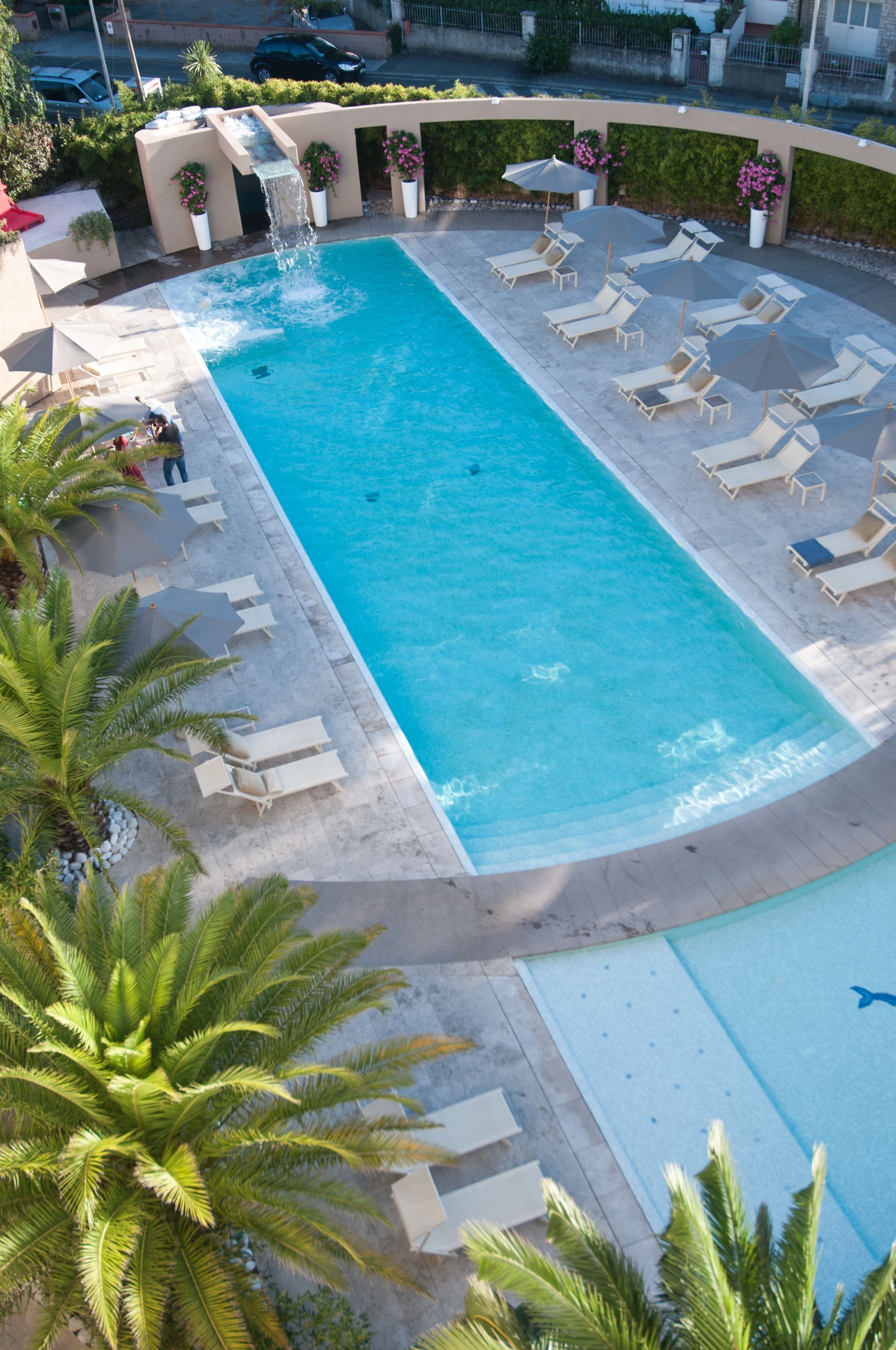 A rectangular swimming pool with a waterfall feature, surrounded by lounge chairs and umbrellas, with palm trees and a paved deck.