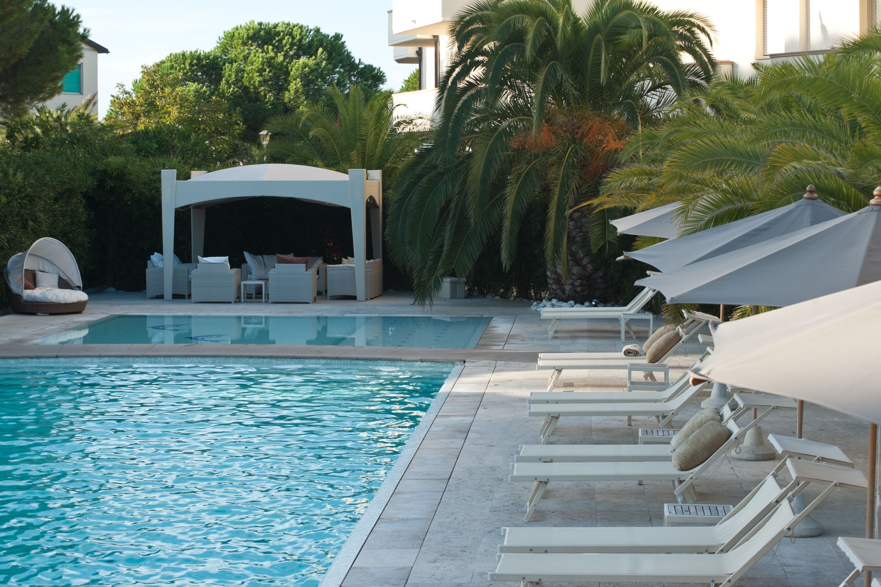 Une piscine entourée de palmiers, avec des chaises longues et des parasols, ainsi qu’un espace de détente abrité.