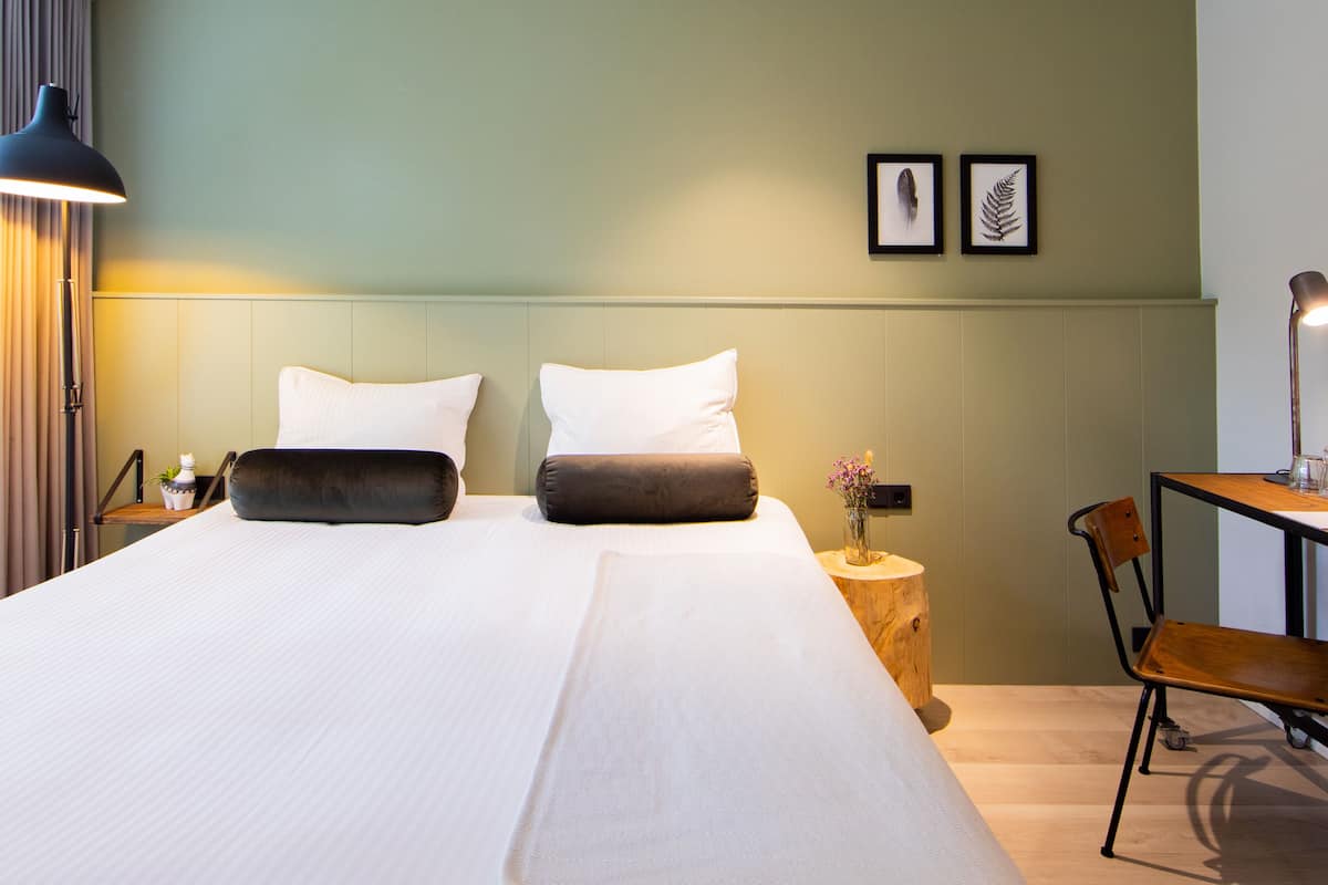 A hotel room with a large bed, two pillows, a bedside table with a lamp, a chair, and a desk with a lamp. There are framed pictures on the wall.