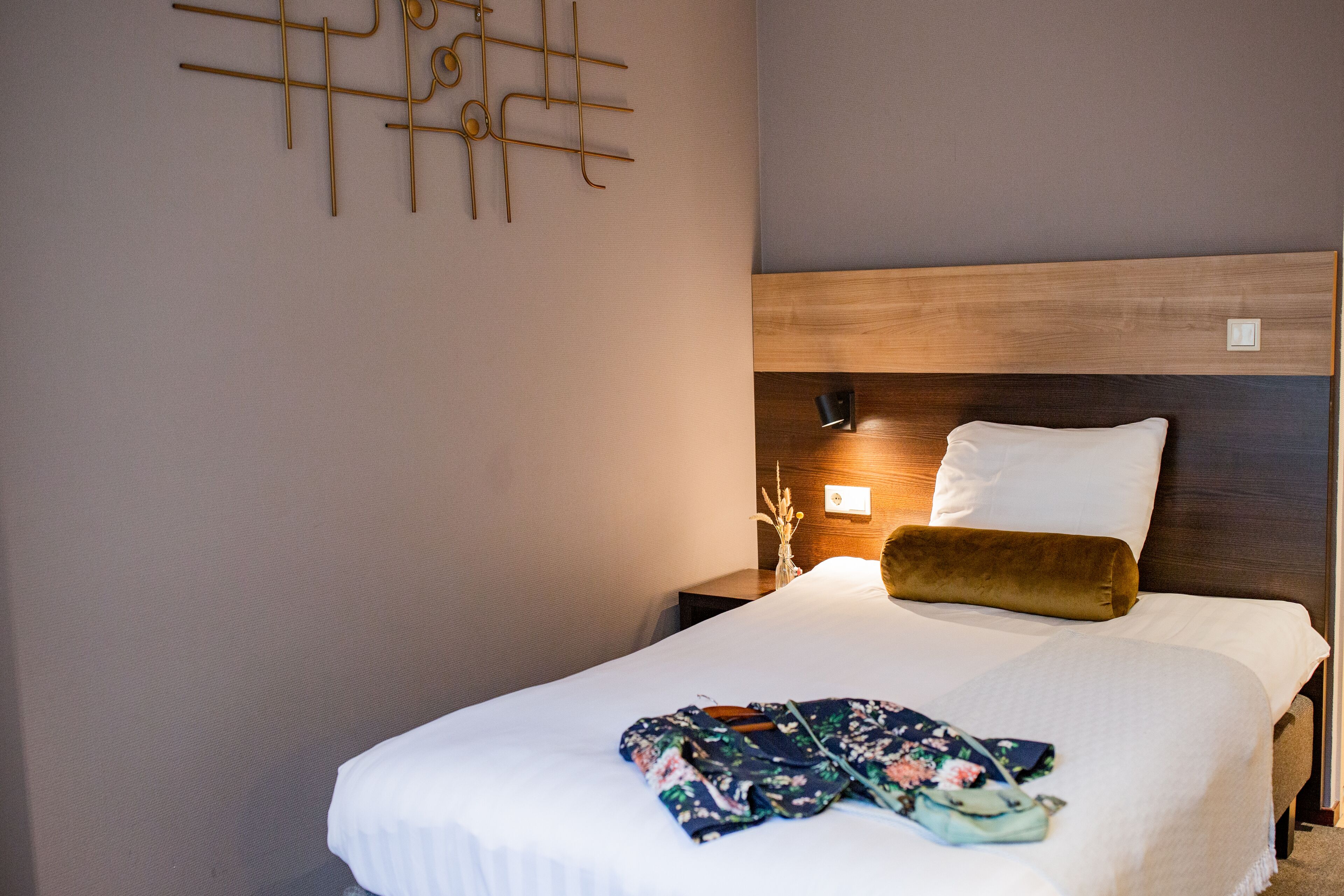 A neatly made bed with a white comforter and a floral blanket, a wooden headboard, a bedside table with a lamp, and a wall art piece.