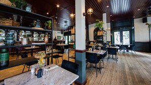 Interior de restaurante con suelos de madera, mesas, sillas y estantes con plantas y botellas.