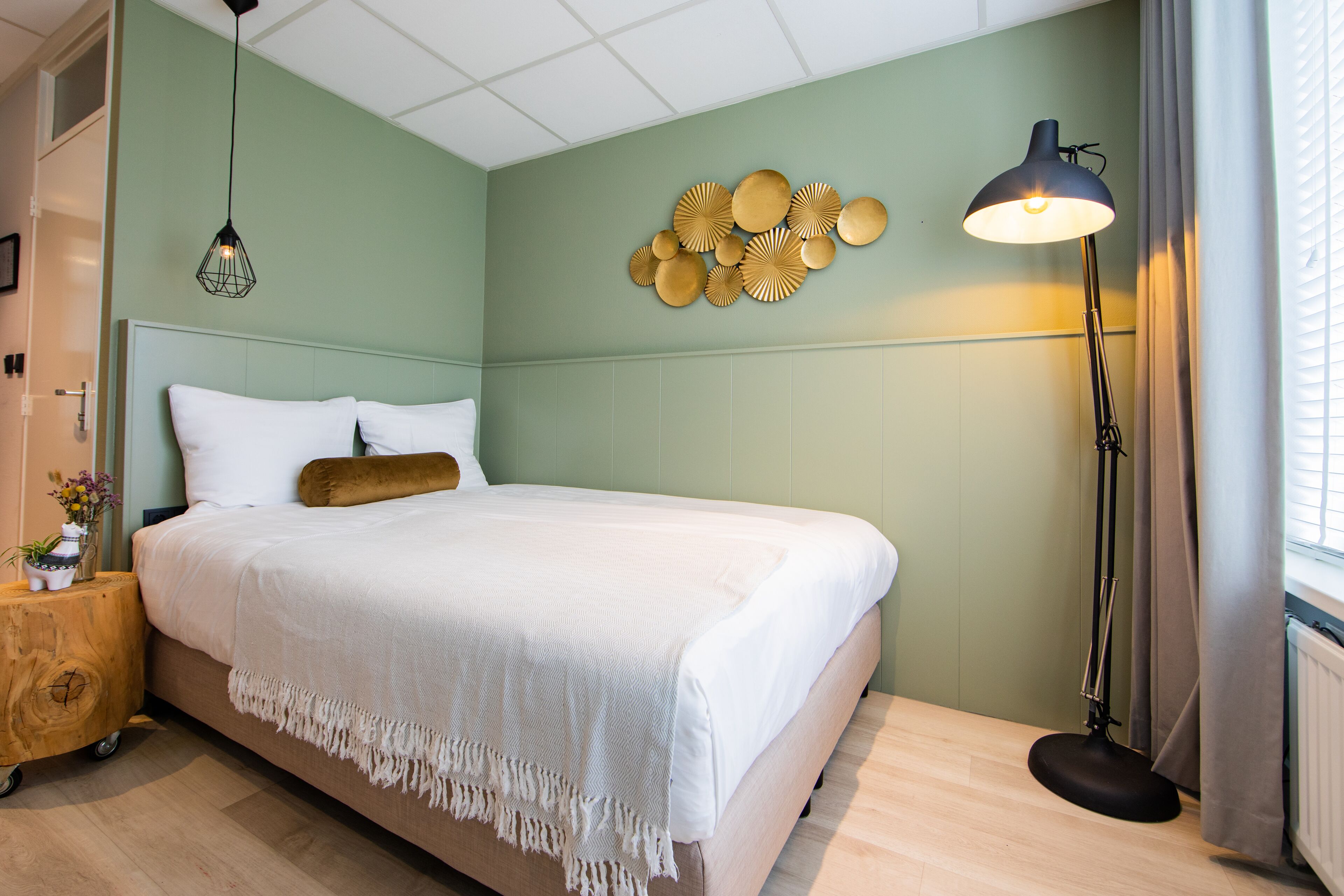 A neatly made bed with a white duvet and a brown pillow, a wooden nightstand with a plant, and a wall-mounted light fixture.
